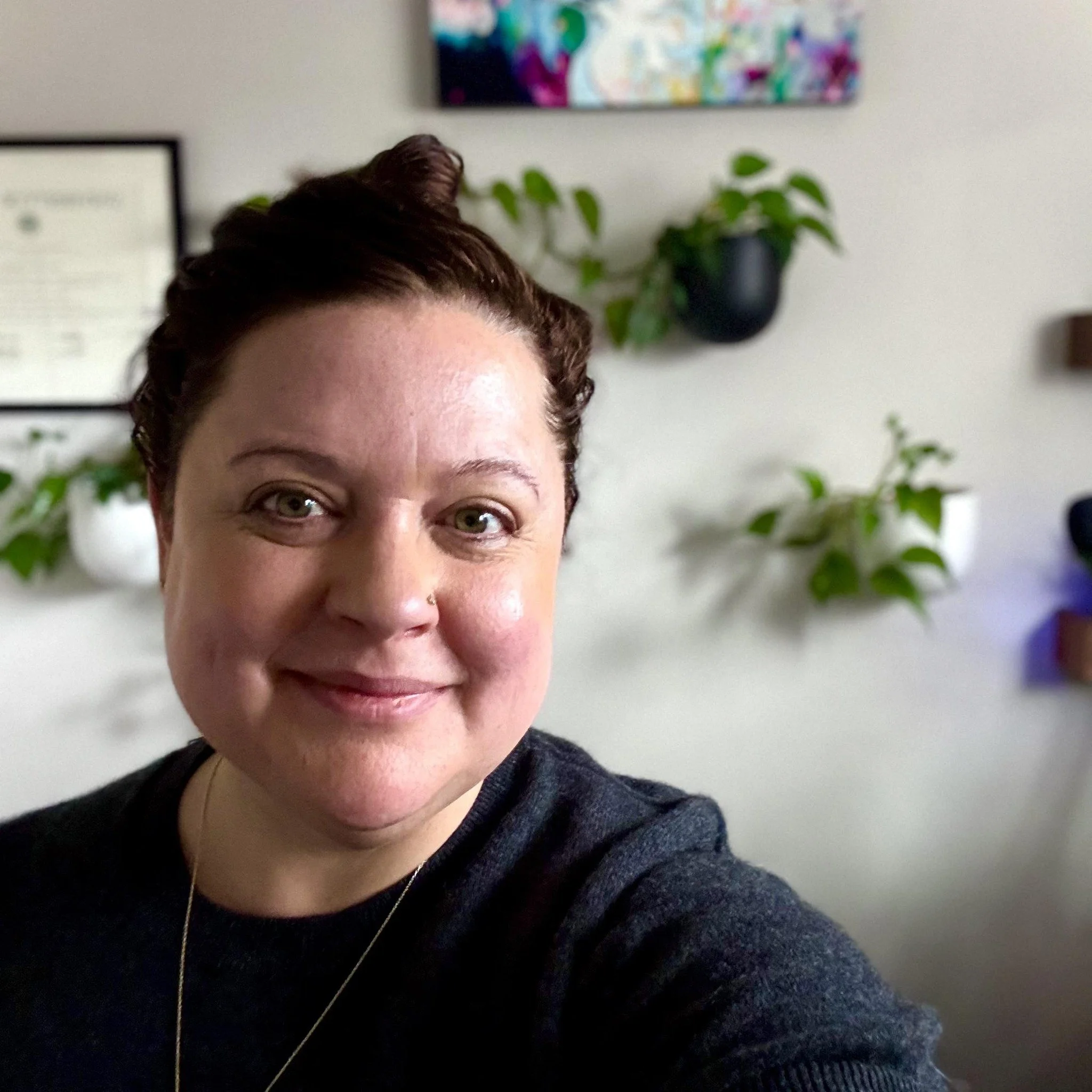 Close-up photo of Meredith, with dark hair in a bun and plants on a wall behind her.