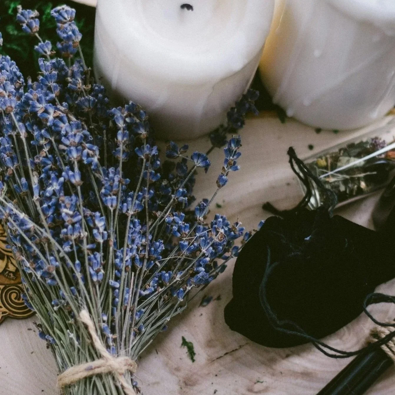 A bundle of dried lavender flowers tied with twine, placed on a wooden surface next to white candles, a black velvet pouch, and a container with dark liquid.