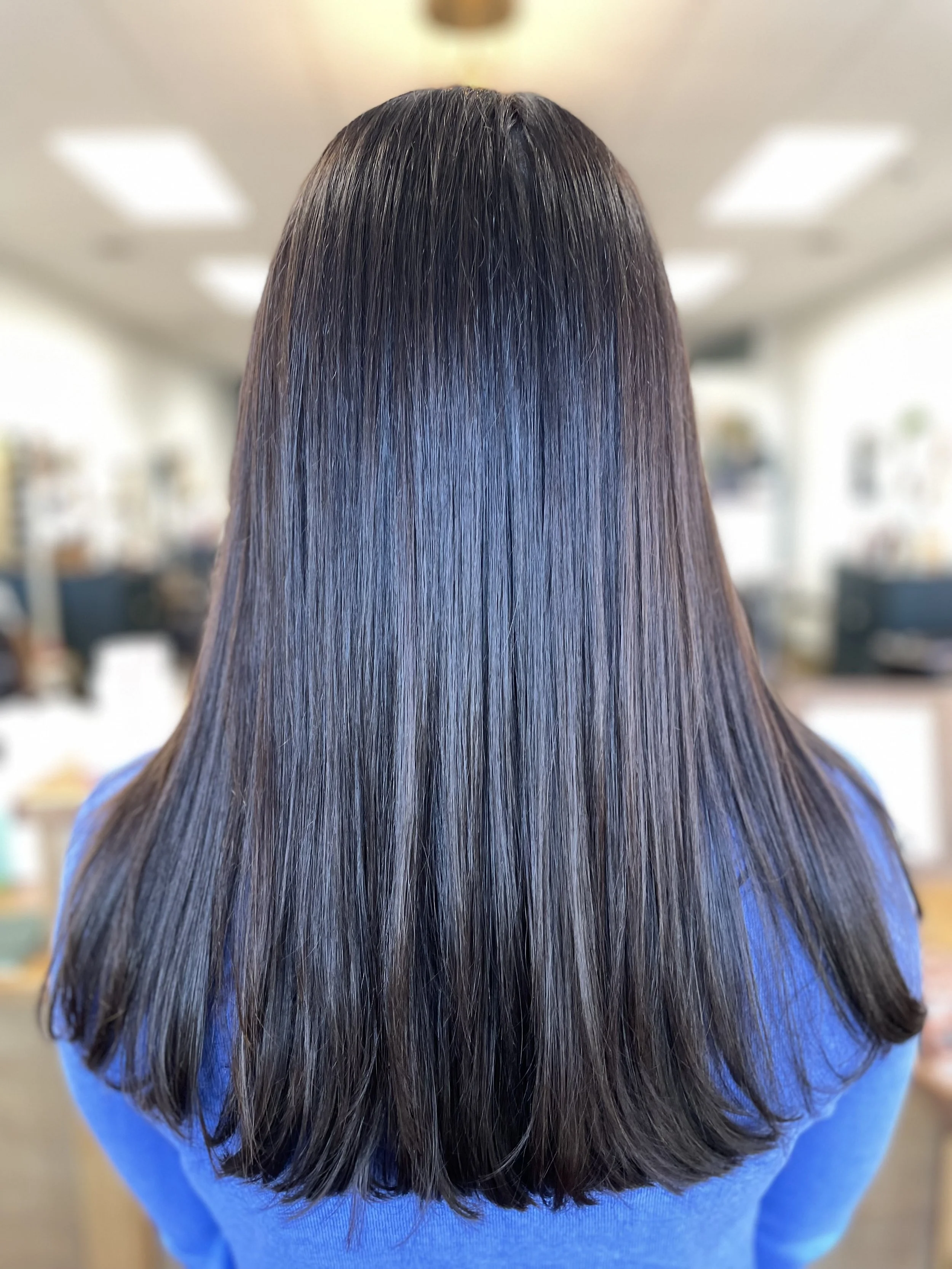 Back view of a woman with long, shiny, dark brown hair with subtle highlights, wearing a blue top, in an indoor setting.