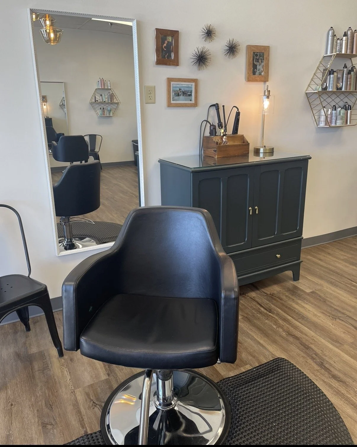 Empty salon chair and mirror reflecting another salon chair, with wall decor and shelves containing hair products.