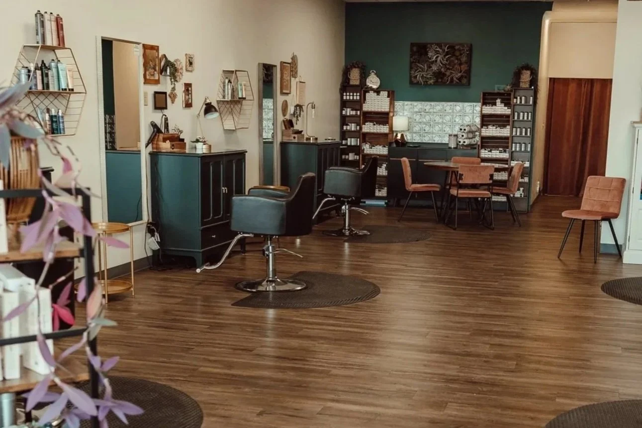 Interior of a hair salon with styling stations, mirrors, and chairs, featuring a combination of modern and vintage decor with wooden flooring and decorative wall shelving.
