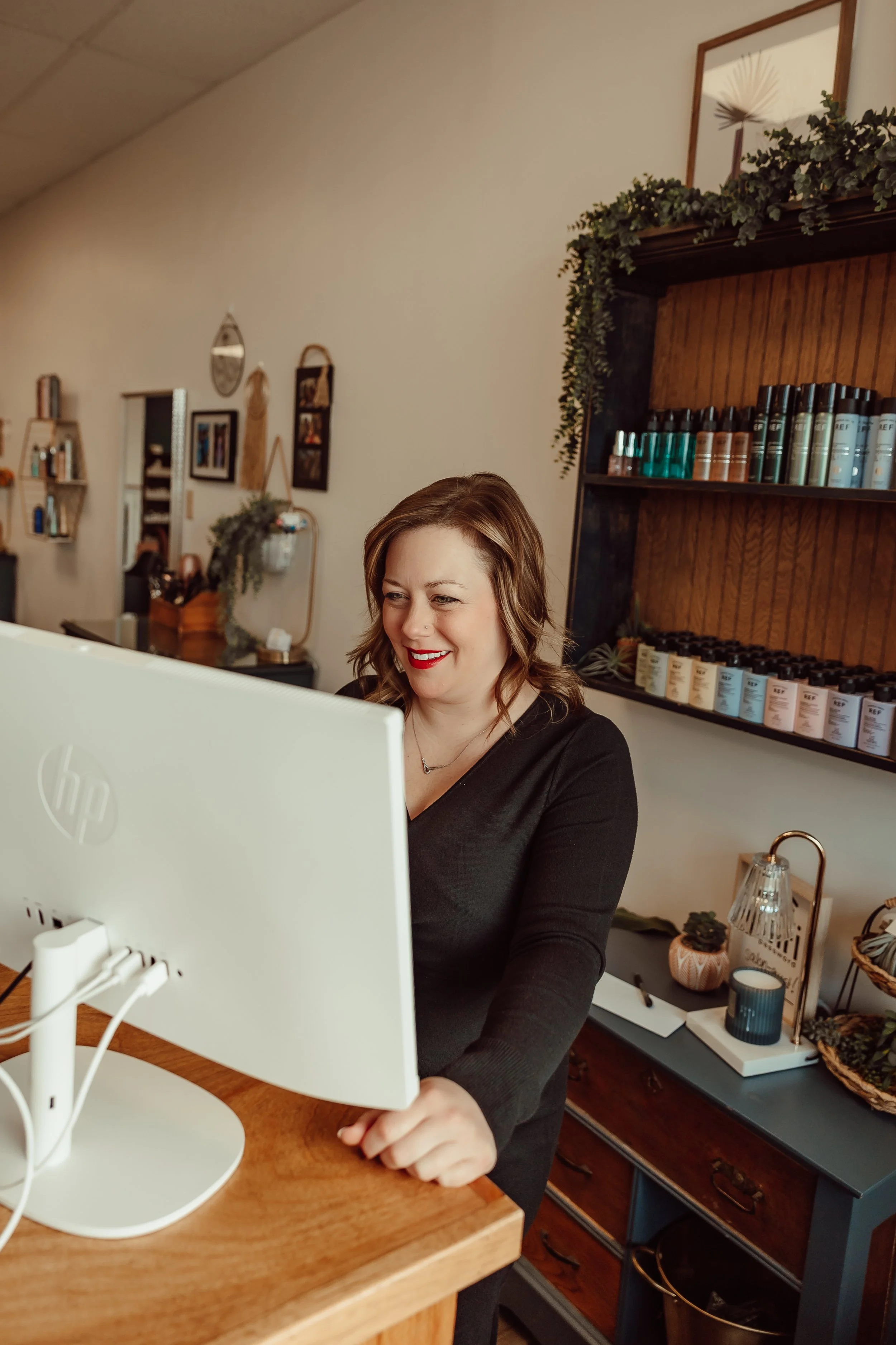A woman with brown hair and red lipstick smiling while working on a computer at a wooden desk in a cozy, decorated space.