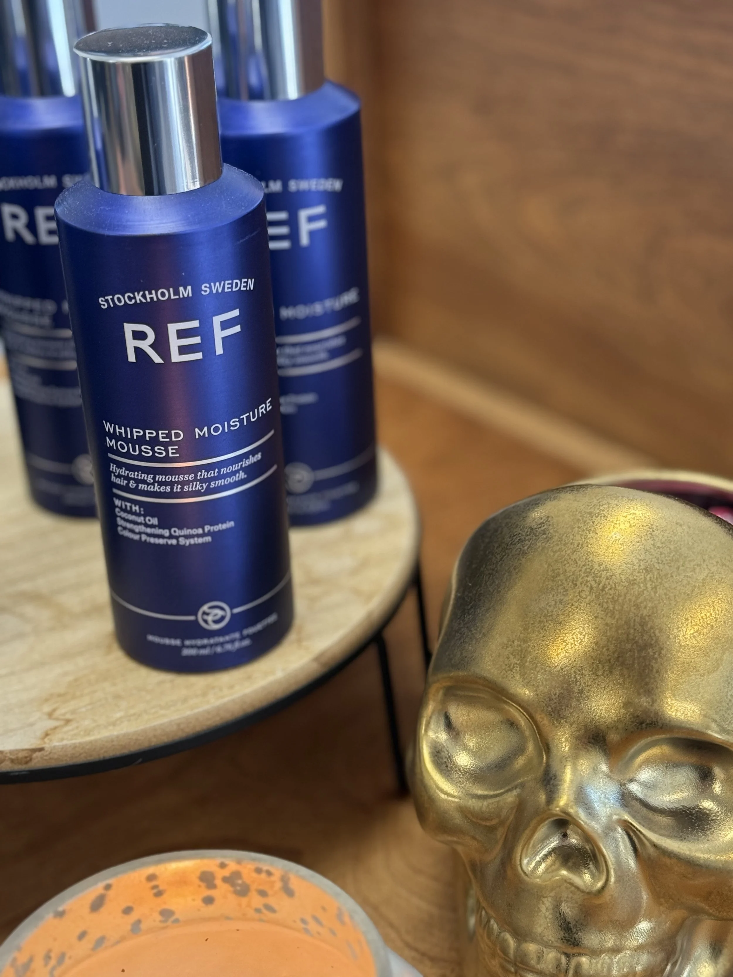 Close-up of blue bottles labeled REF Whipped Moisture Mousse on a wooden surface, next to a gold skull decorative item, with a candle in the foreground.