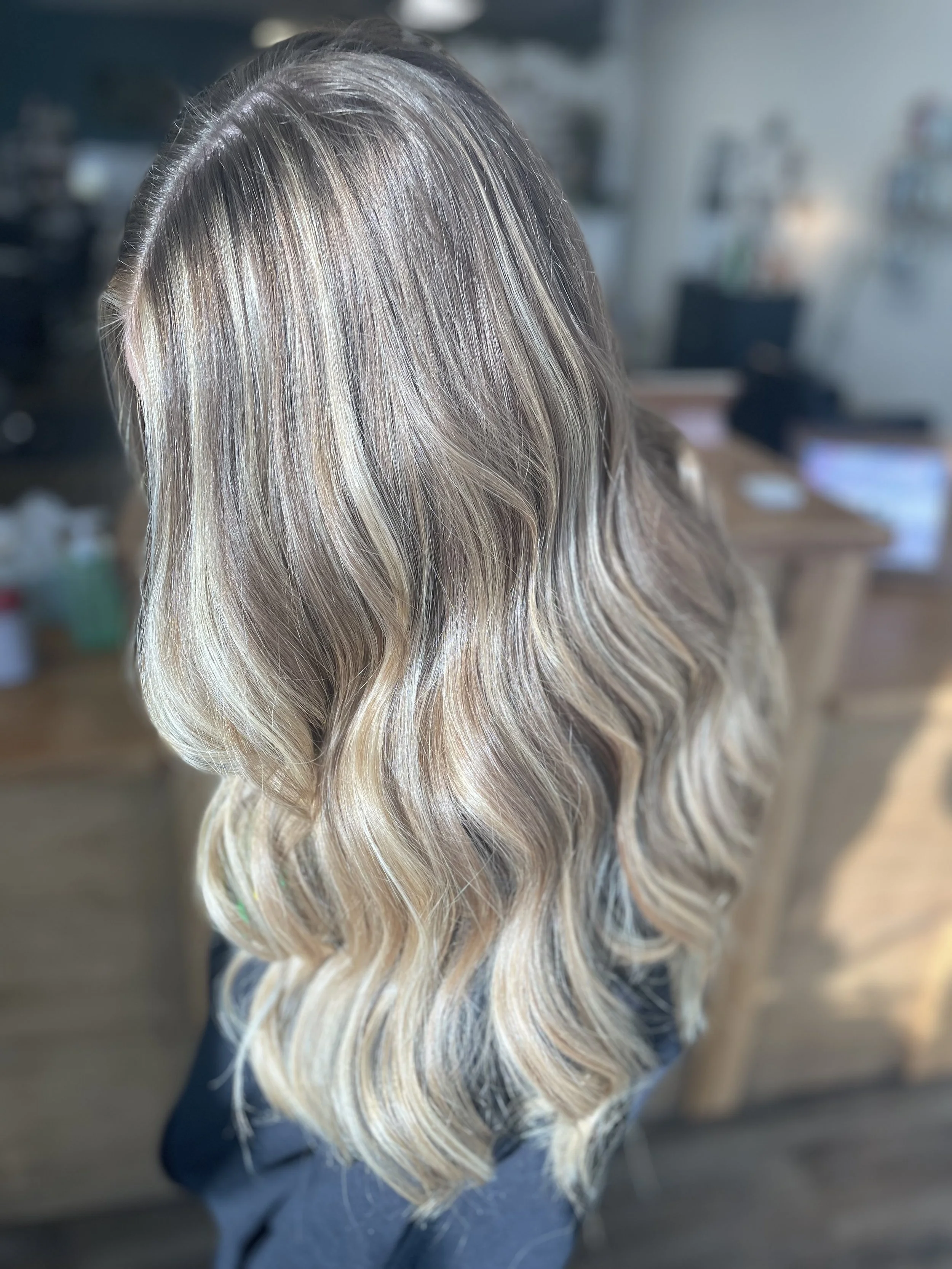 Close-up of a woman's wavy, shoulder-length hair with blonde highlights and darker roots, in a salon setting.
