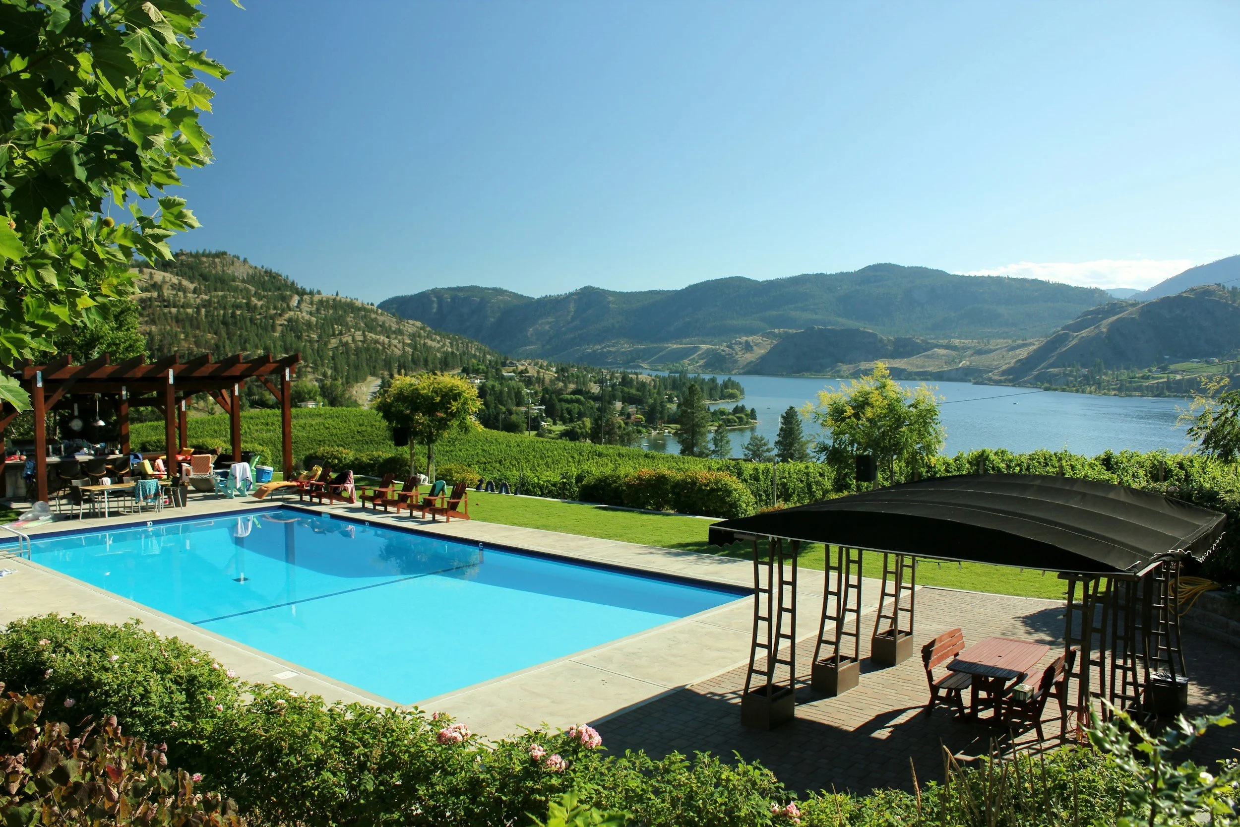 Outdoor swimming pool with lounge chairs, shaded sitting area, set amid lush green landscape with trees and a scenic lake and mountains in the background on a sunny day.