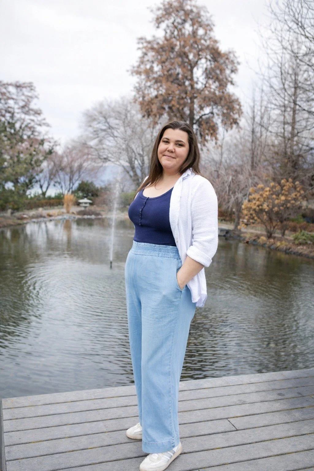 Young woman standing on a wooden dock near a pond with trees in the background, wearing a white cardigan, navy top, light blue pants, and white sneakers.