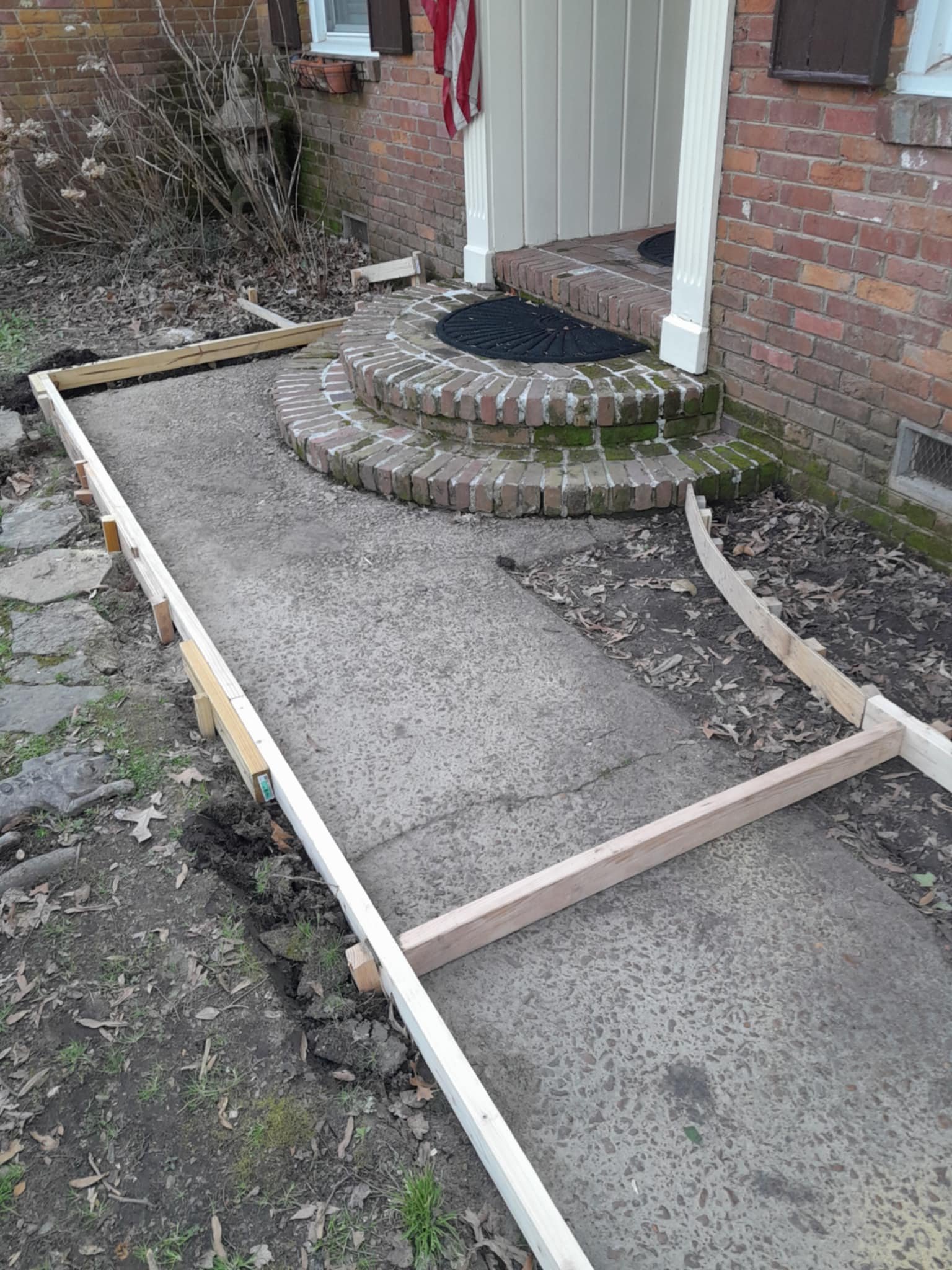 Construction work in progress on a new sidewalk in front of a brick house, with a wooden frame outlining the area to be paved.