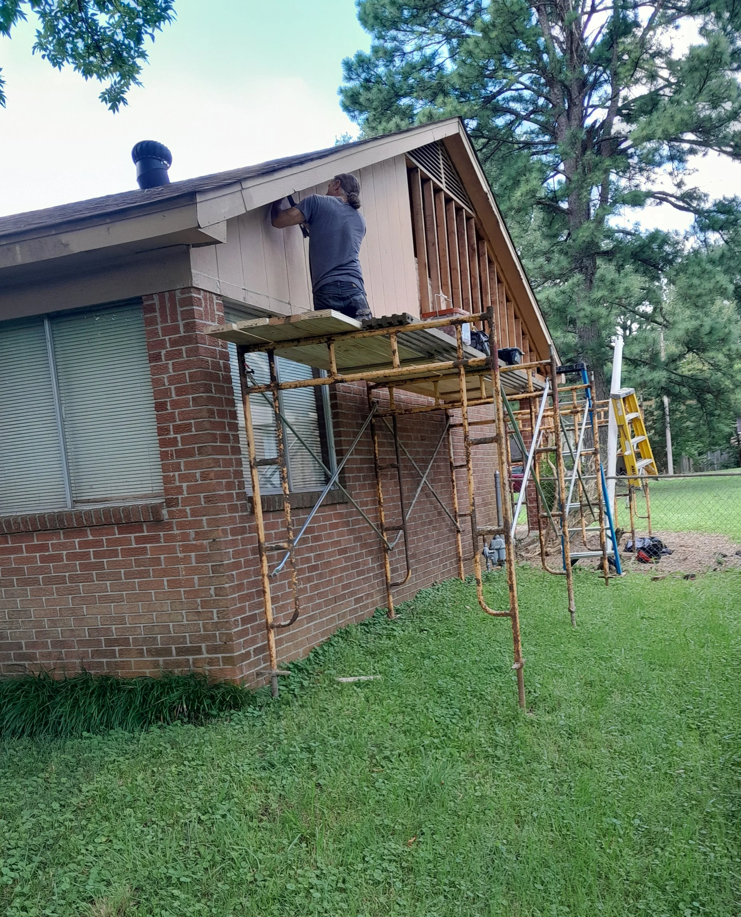 A person working on the exterior of a house, standing on scaffolding, installing or repairing the siding under the roof eaves, with trees visible in the background.