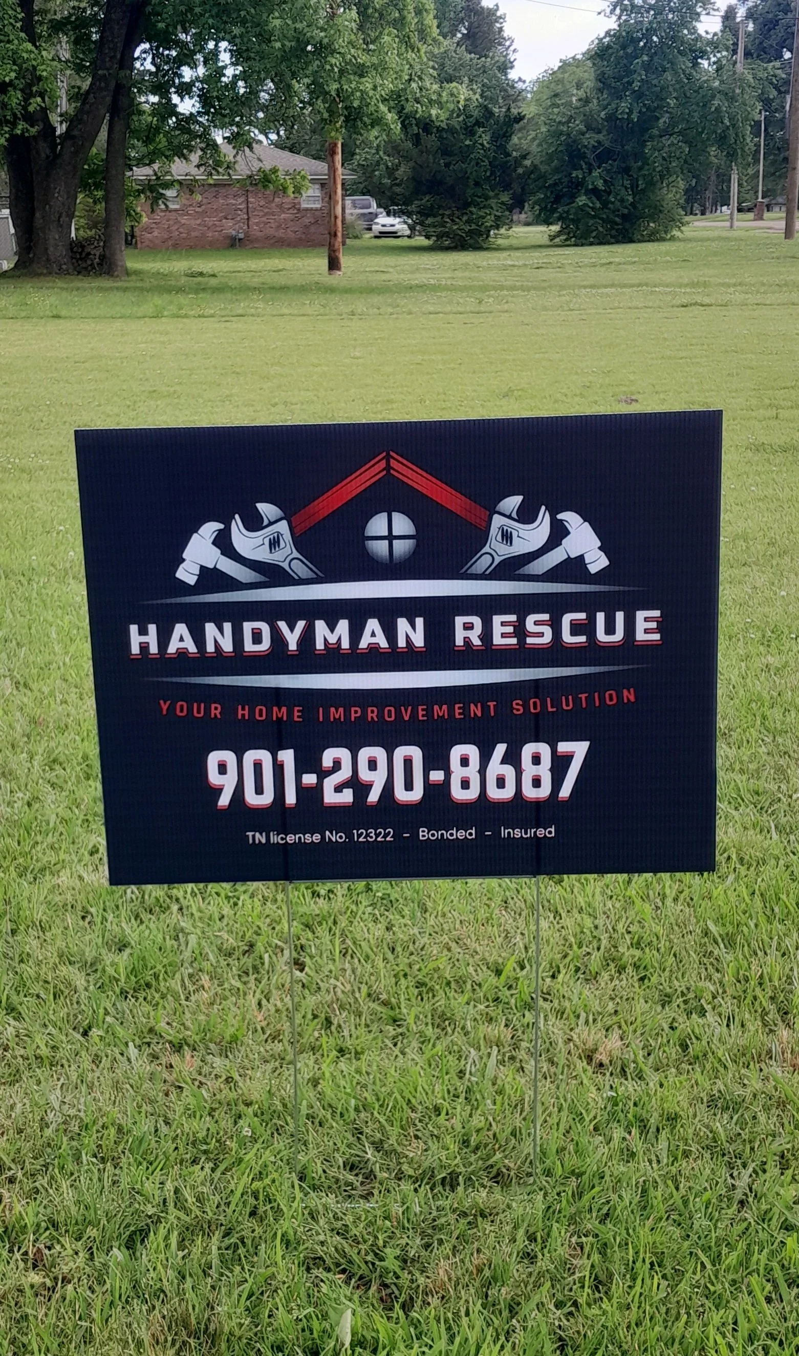 Sign for Handyman Rescue services on a grassy lawn, featuring tools and a house at the top, with contact information and licensing details.