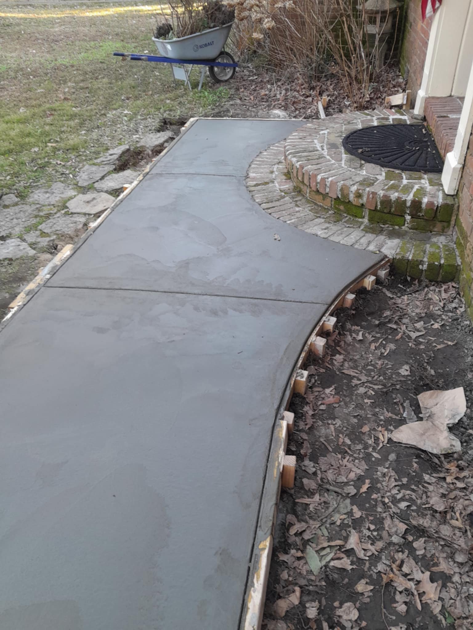 Newly poured concrete walkway with wooden forms along the edges, leading to a brick porch with a curved brick step. A black round door mat is on the porch, and a gray wheelbarrow is in the background on the grass.