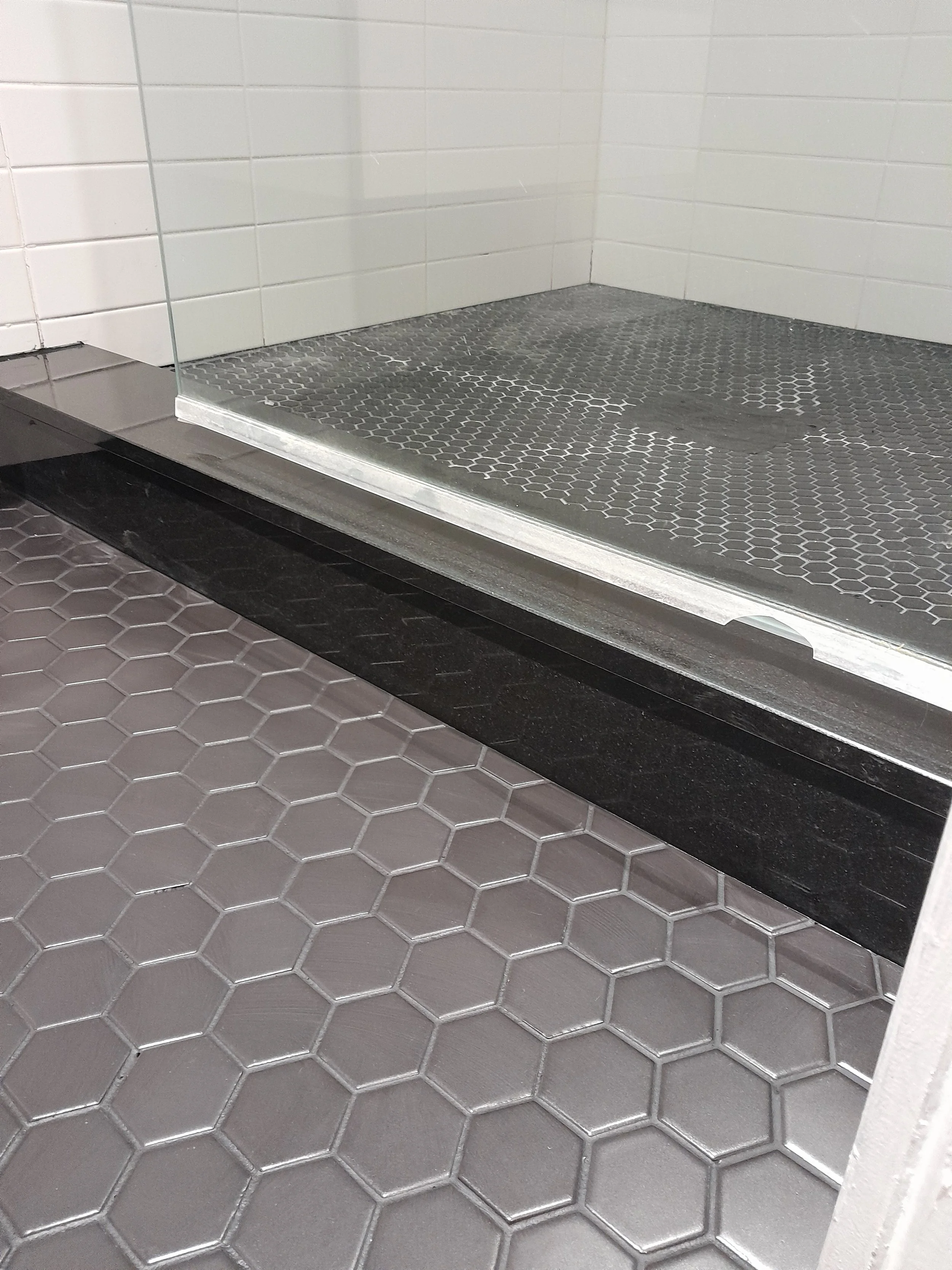 A shower with black hexagonal floor tiles, a glass partition, and white tiled walls.