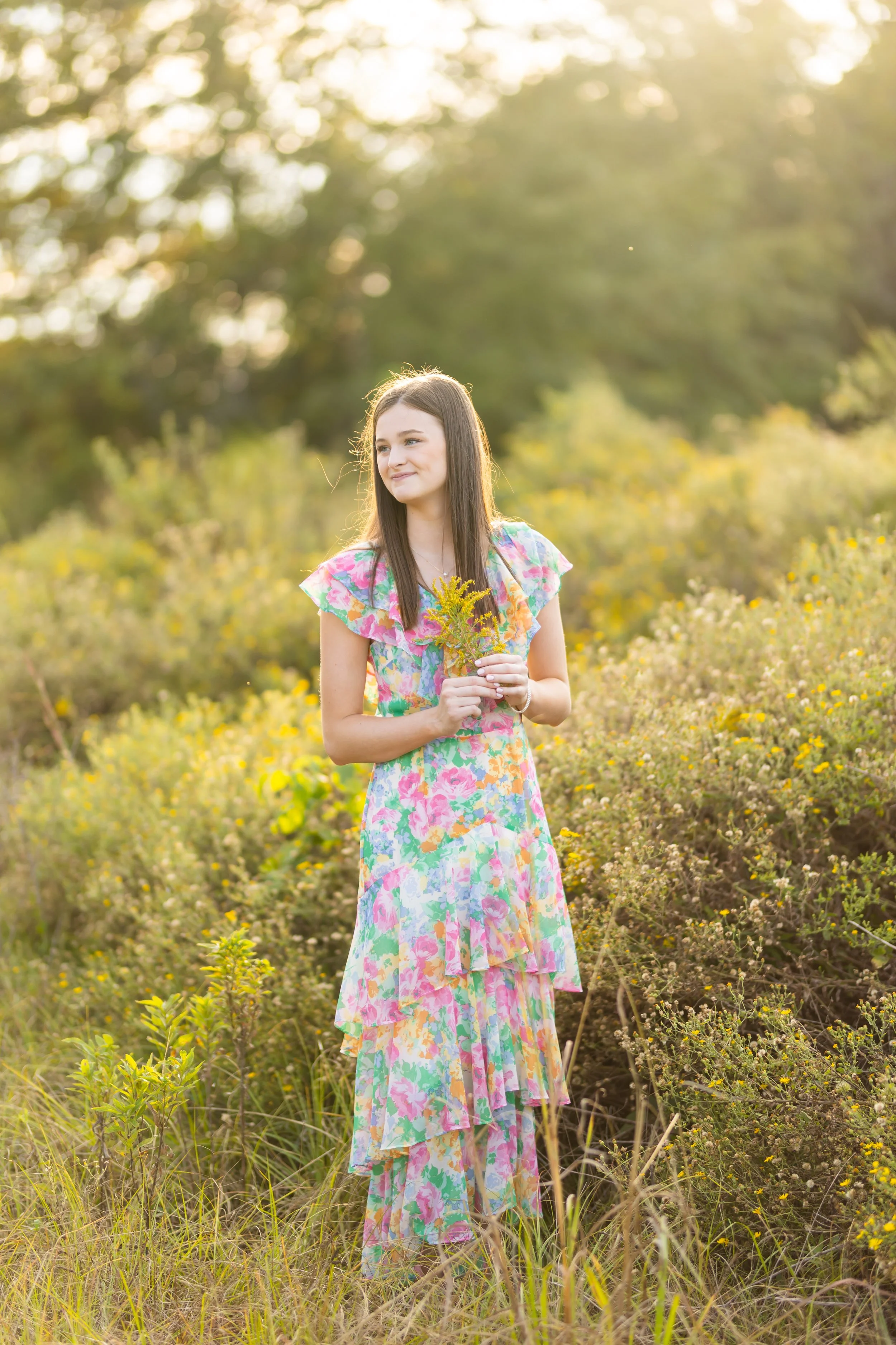 Ruth in a colorful, floral dress standing in a field of yellow wildflowers, holding a small bouquet, with a soft, golden sunset in the background.