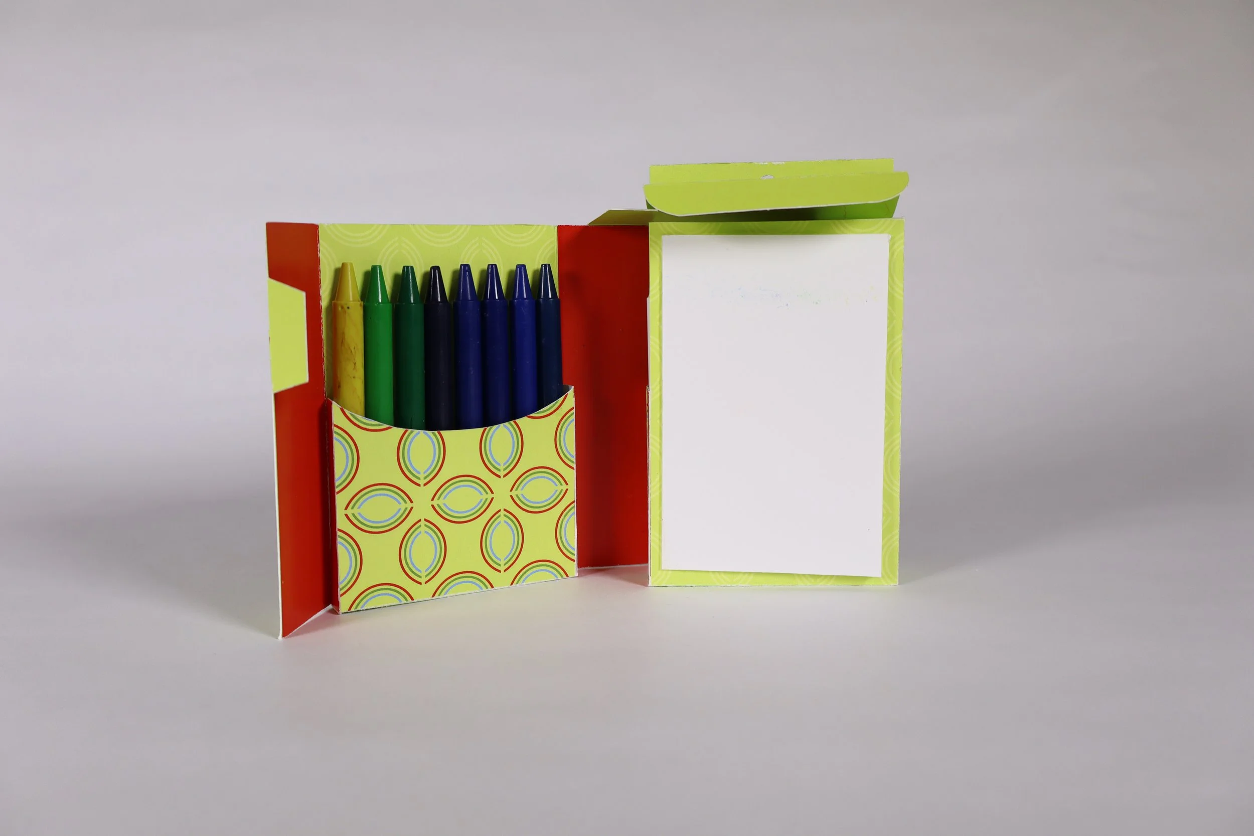 Open redesigned crayon box displaying green and blue crayons in a patterned pocket beside a blank white paper panel.