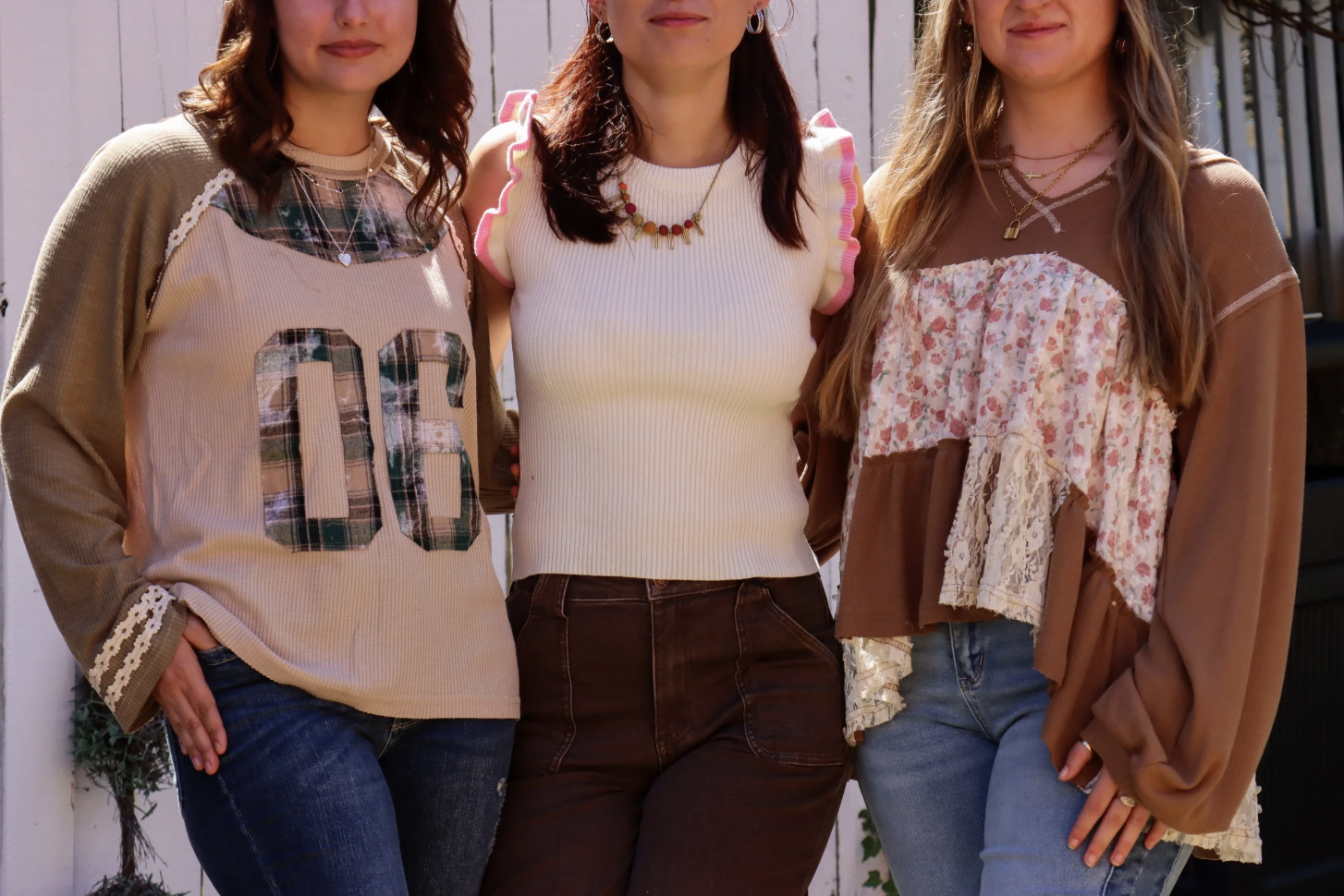 Close-up of three women standing outdoors in front of a white fence, shown from the shoulders to hips, with one wearing a beige shirt with plaid and large number 06, the second wearing a sleeveless white top with pink ruffle sleeves, and the third we