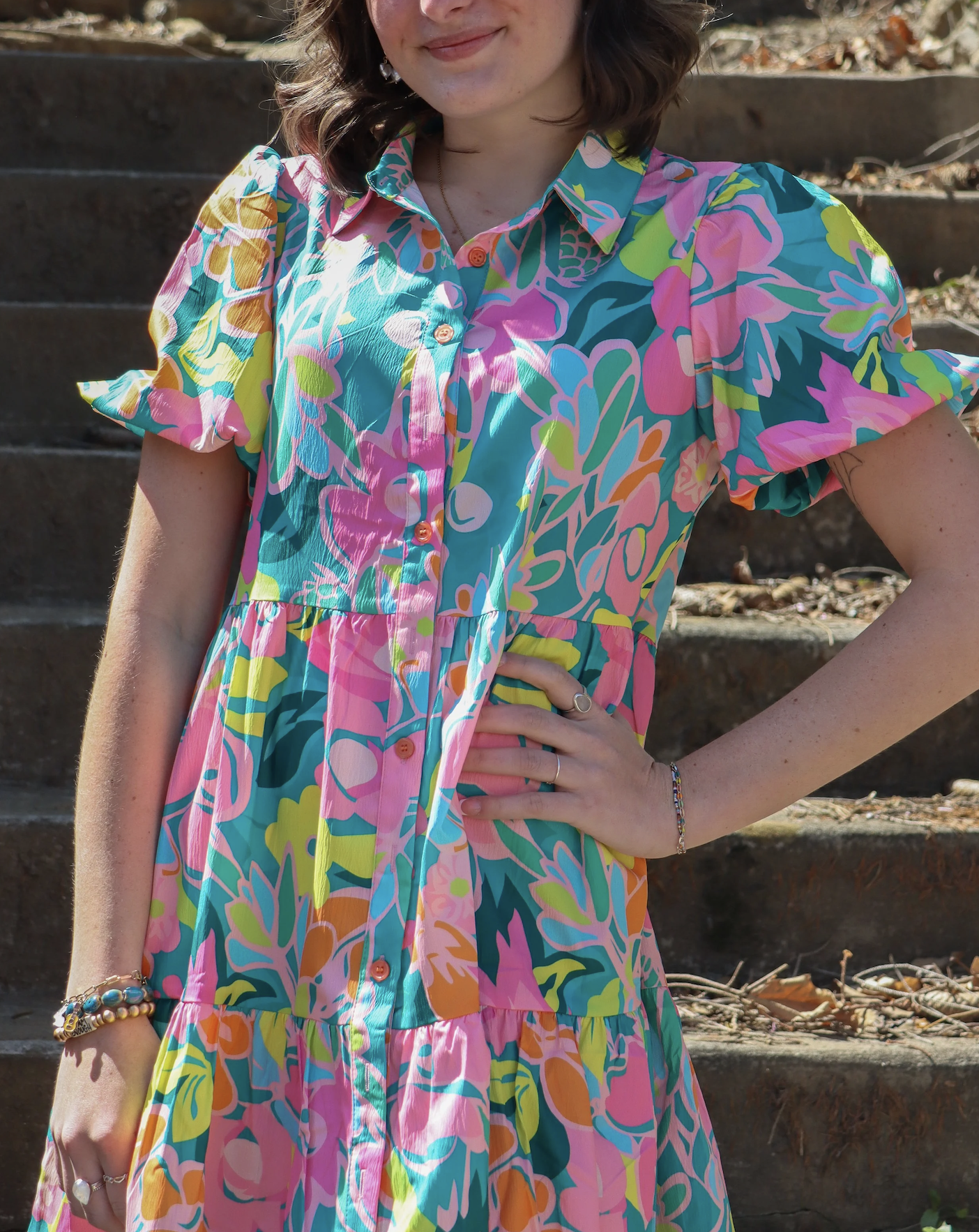 A woman wearing a colorful, floral-patterned dress with short sleeves, standing on outdoor stairs.