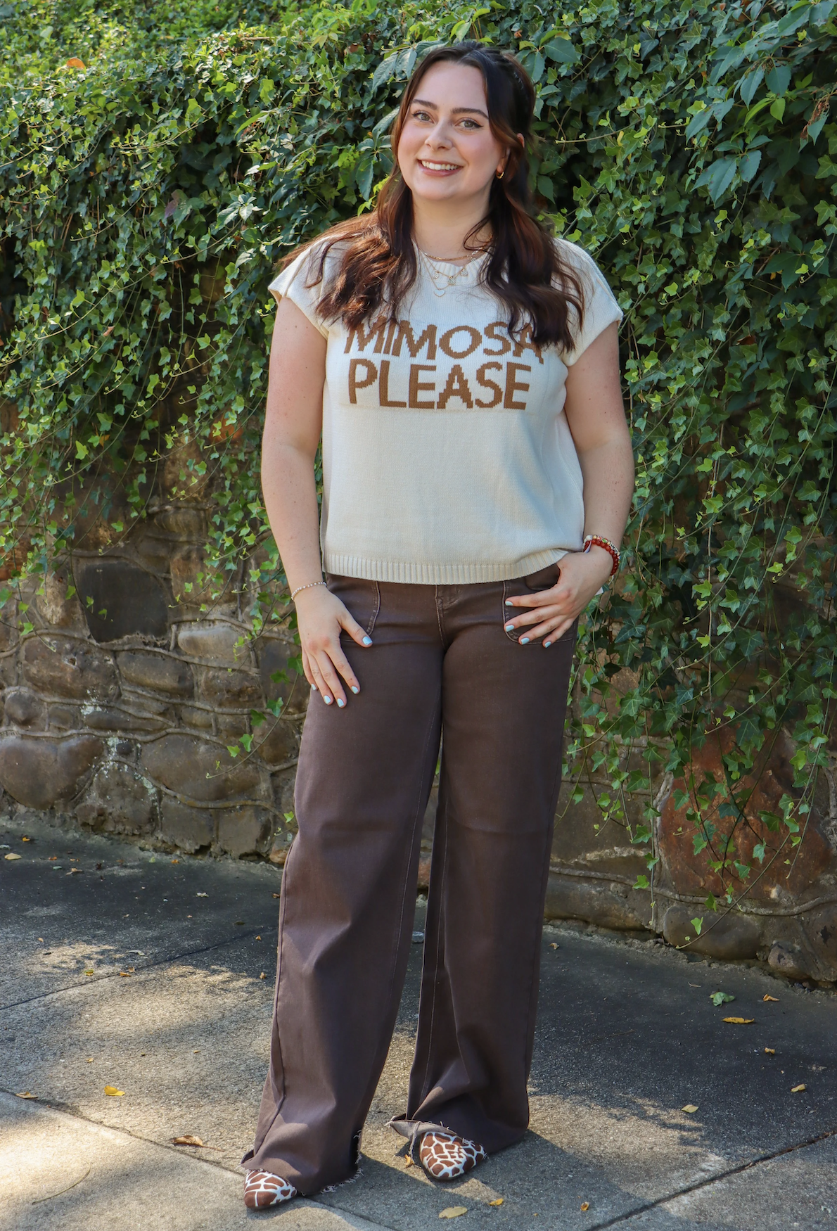 A young woman with dark brown hair and light skin standing outdoors in front of a green leafy background. She is smiling, wearing a light-colored sweater with the words 'MIMOSA PLEASE' written on it, brown wide-leg pants, and animal print shoes.