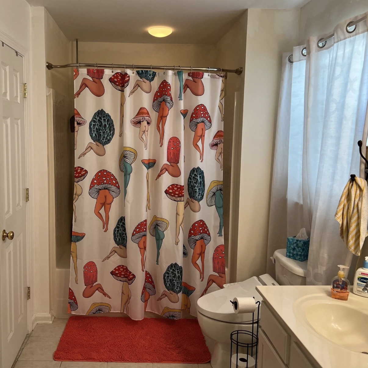 A bathroom with a shower curtain featuring cartoon legs and mushroom heads, a red bath mat, and bathroom toiletries on the sink counter.