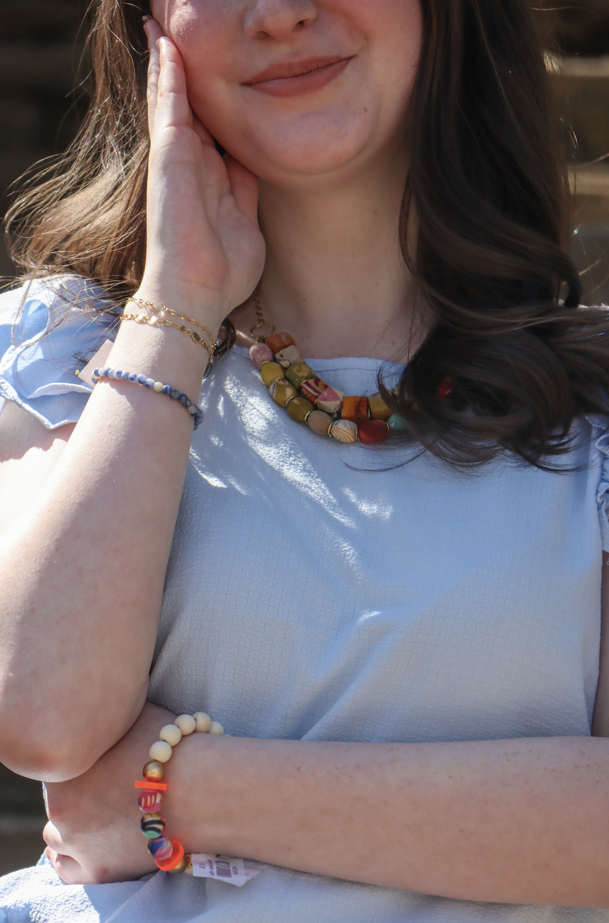 Close-up of a woman wearing a light blue top, colorful beaded necklace, and various bracelets, with sunlit hair and a gentle smile.