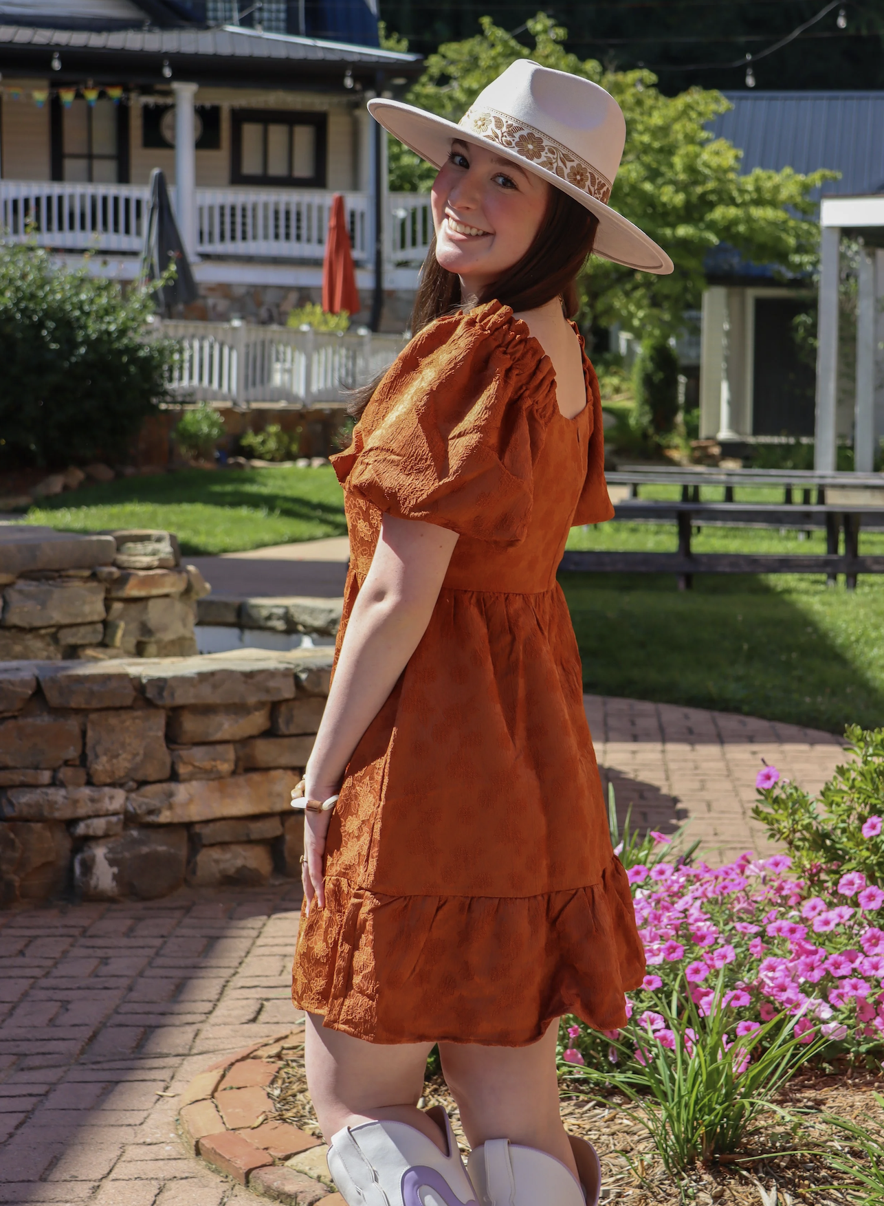 Young woman in an orange dress with puffed sleeves and white cowboy boots, smiling and looking over her shoulder outdoors in a garden with pink flowers and greenery, wearing a wide-brimmed hat with a floral band.