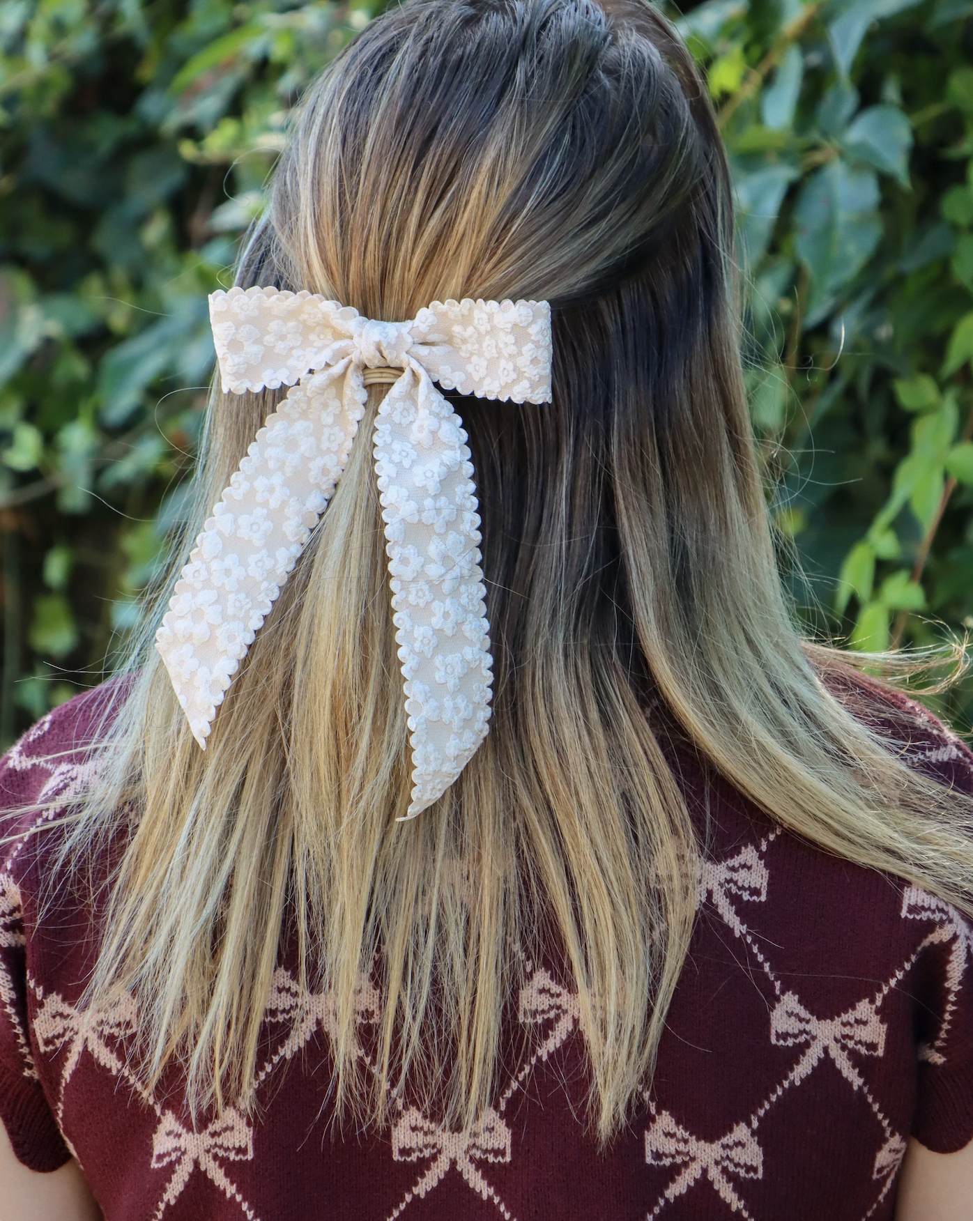 Back of a woman's head with blonde hair tied with a white floral-patterned lace bow, outdoors with green foliage in the background.