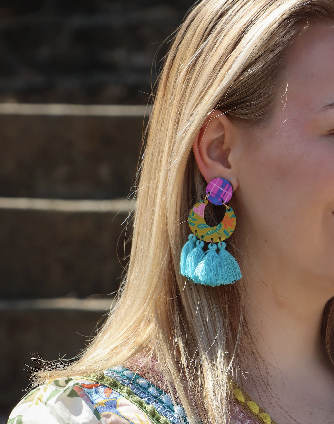 Close-up of a woman wearing colorful, patterned earrings with turquoise tassels, showing her blonde hair and part of her face against a dark background.