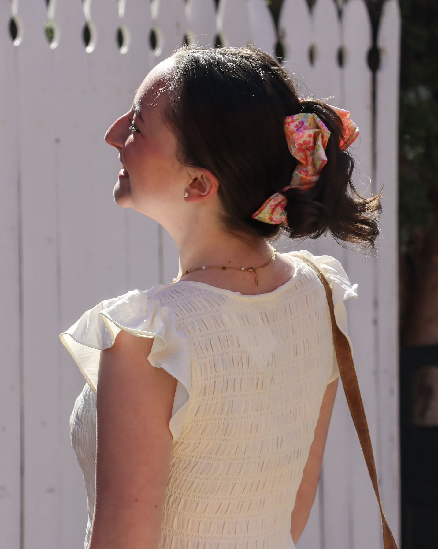 Profile of a woman with dark hair in a bun, wearing a floral hair scrunchie, a white ruffled top, and a gold necklace, standing outdoors near a white picket fence.