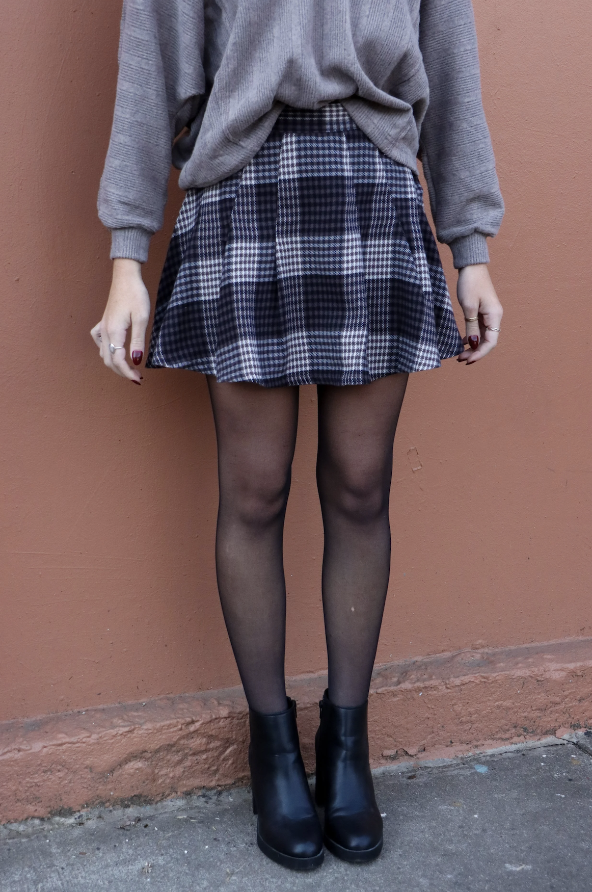 Person wearing a grey sweater, plaid skirt, sheer black tights, and black ankle boots standing against an orange wall.