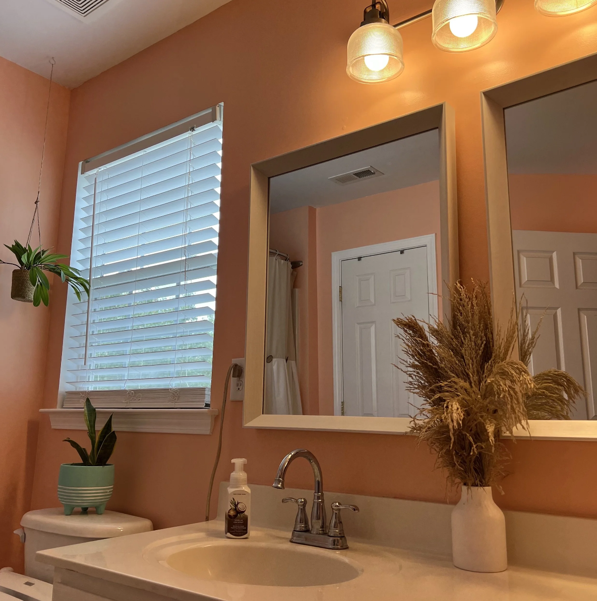 Bathroom with peach-colored walls, a white sink with a silver faucet, a large mirror, a window with white blinds, a potted plant on the toilet tank, a bottle of hand soap, and a vase with dried plants, with warm lighting from three ceiling lights.