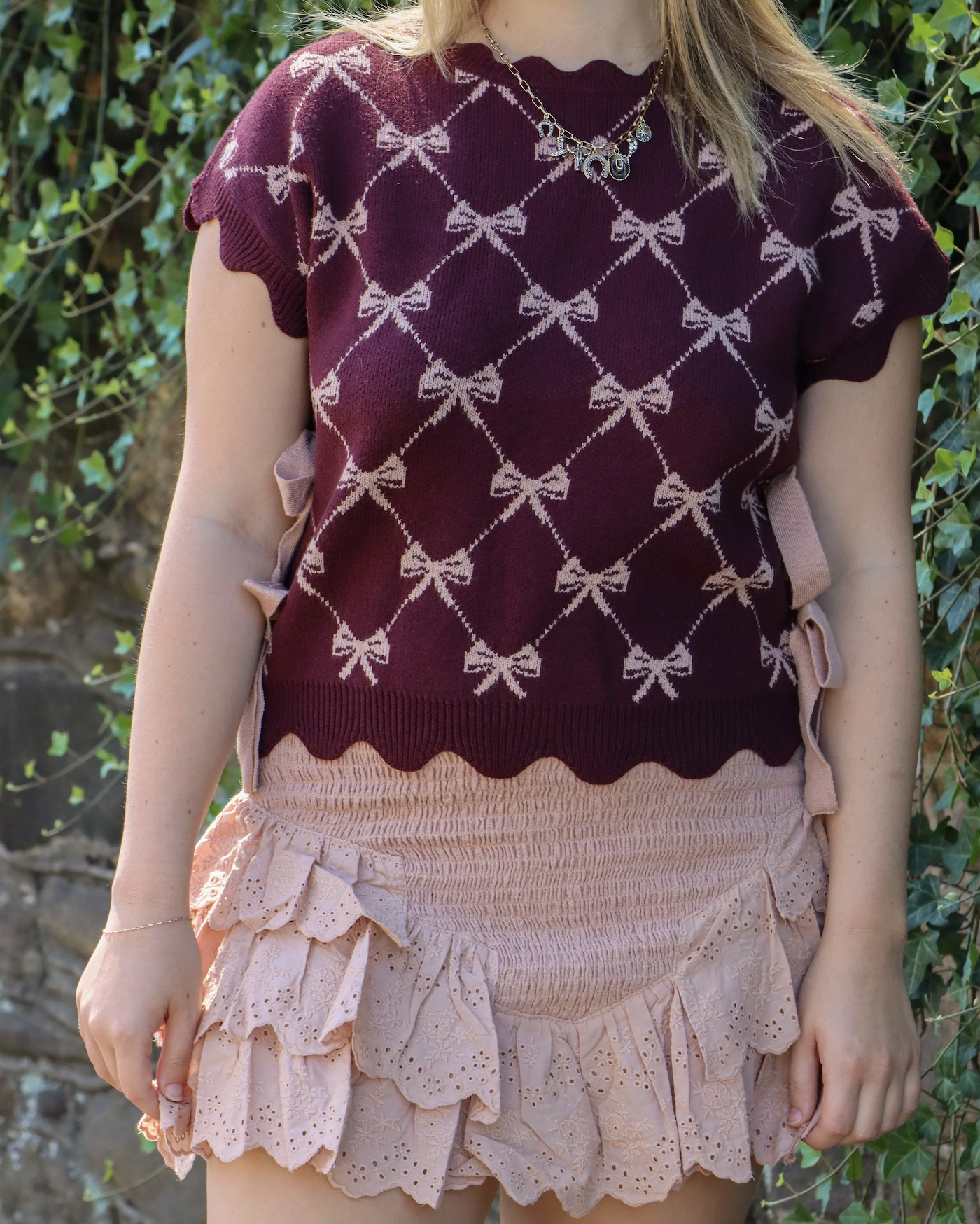 Close-up of a woman wearing a burgundy knitted top with a bow pattern and ruffled sleeves, paired with pink eyelet shorts with ruffles and bow accents, outdoors with greenery in the background.