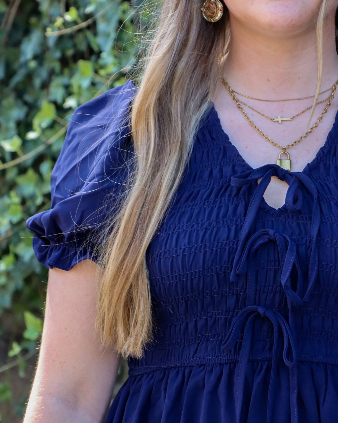 Close-up of a woman wearing layered gold necklaces, gold earrings, and a navy blue dress with a tied neckline, outdoors with greenery in the background.