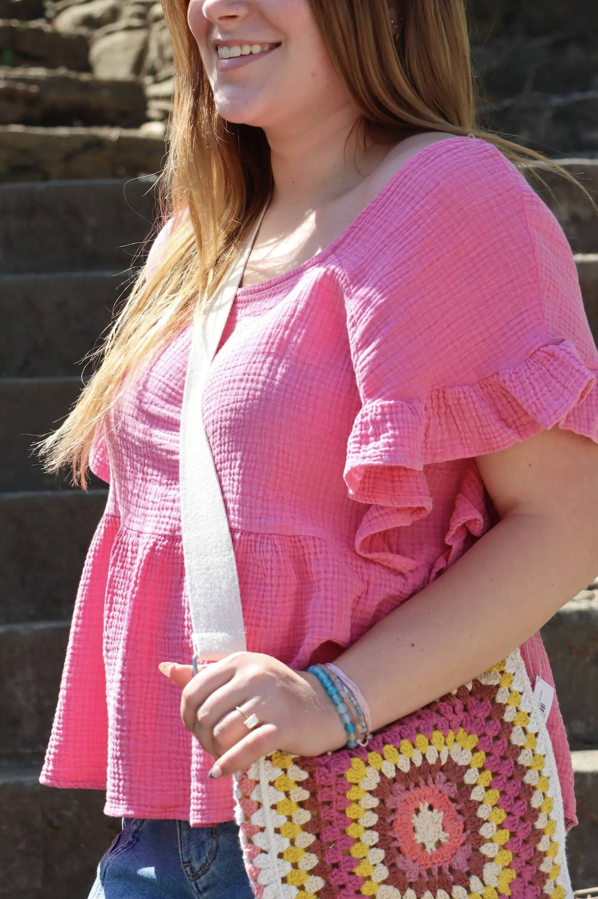A woman with long red hair is smiling, wearing a pink textured blouse with ruffled sleeves, and carrying a colorful crocheted bag with a pink, white, yellow, and orange pattern. She is standing outside on stone steps.