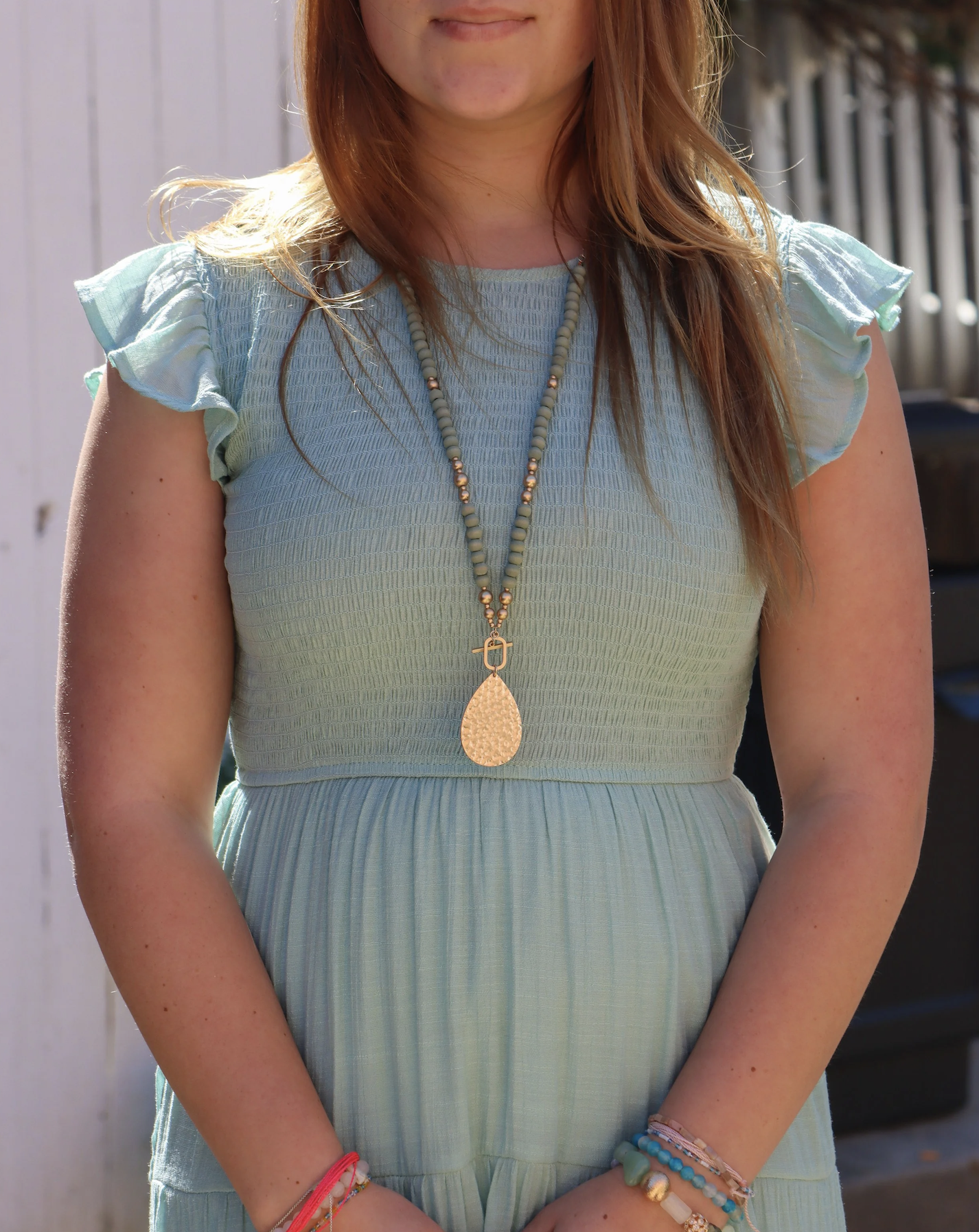 A woman is wearing a light blue dress with ruffled sleeves and a beaded necklace with a large teardrop-shaped pendant, standing outdoors.