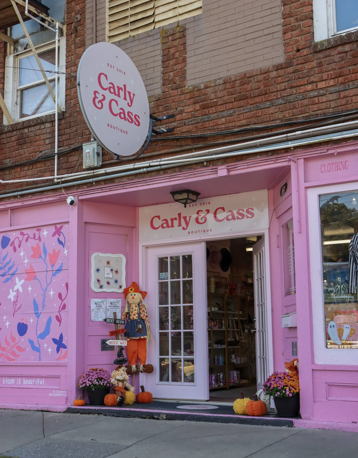 Pink storefront of Carly & Cass boutique with Halloween decorations, pumpkins, and flowers outside, and a sign overhead reading 'Carly & Cass' in red letters.