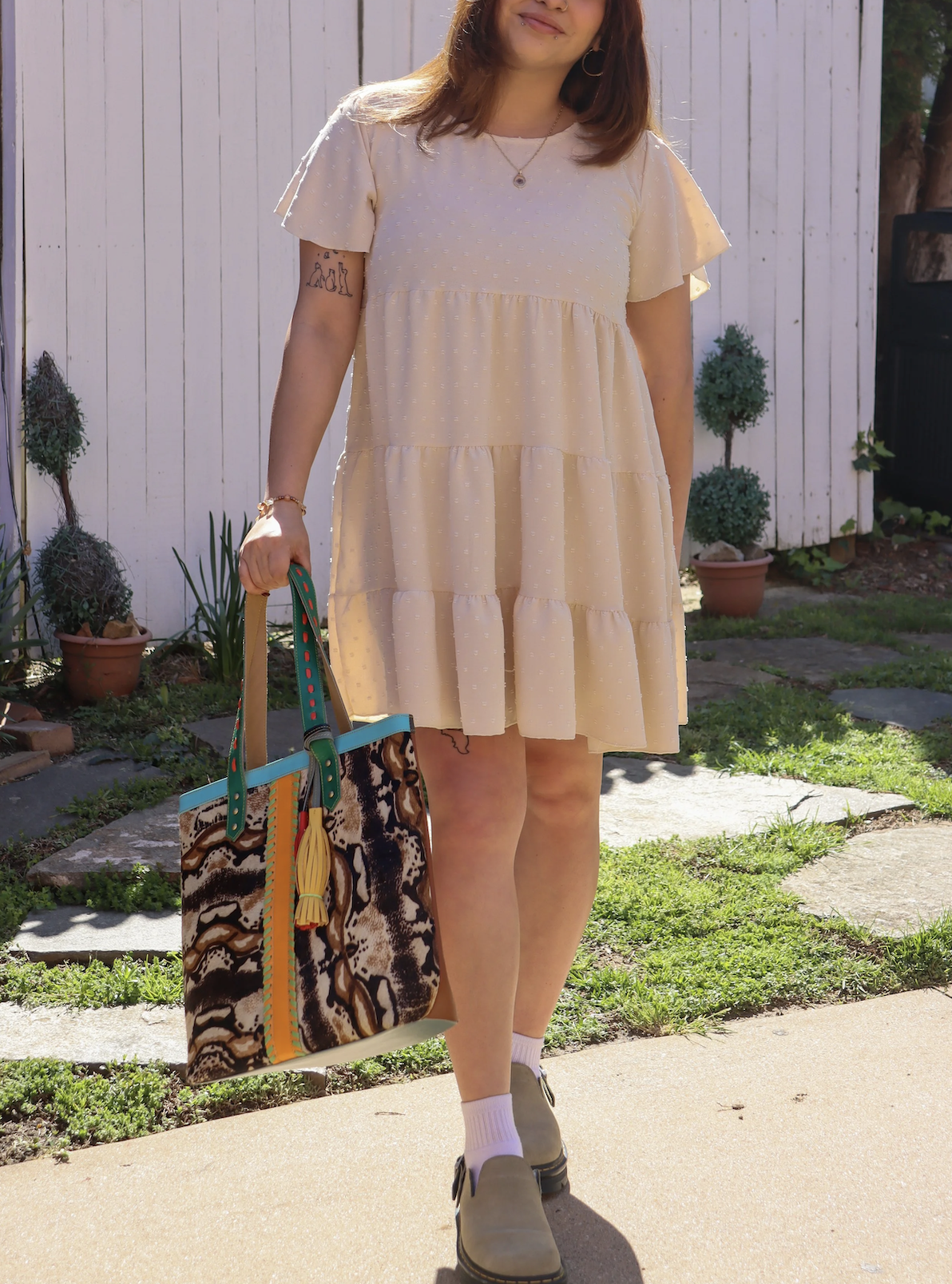 A woman wearing a light beige, tiered dress with puffed sleeves, standing outdoors on a sunny day. She is holding a large, colorful tote bag with a snake pattern and multicolored accents. Potted plants and a white fence are visible in the background.