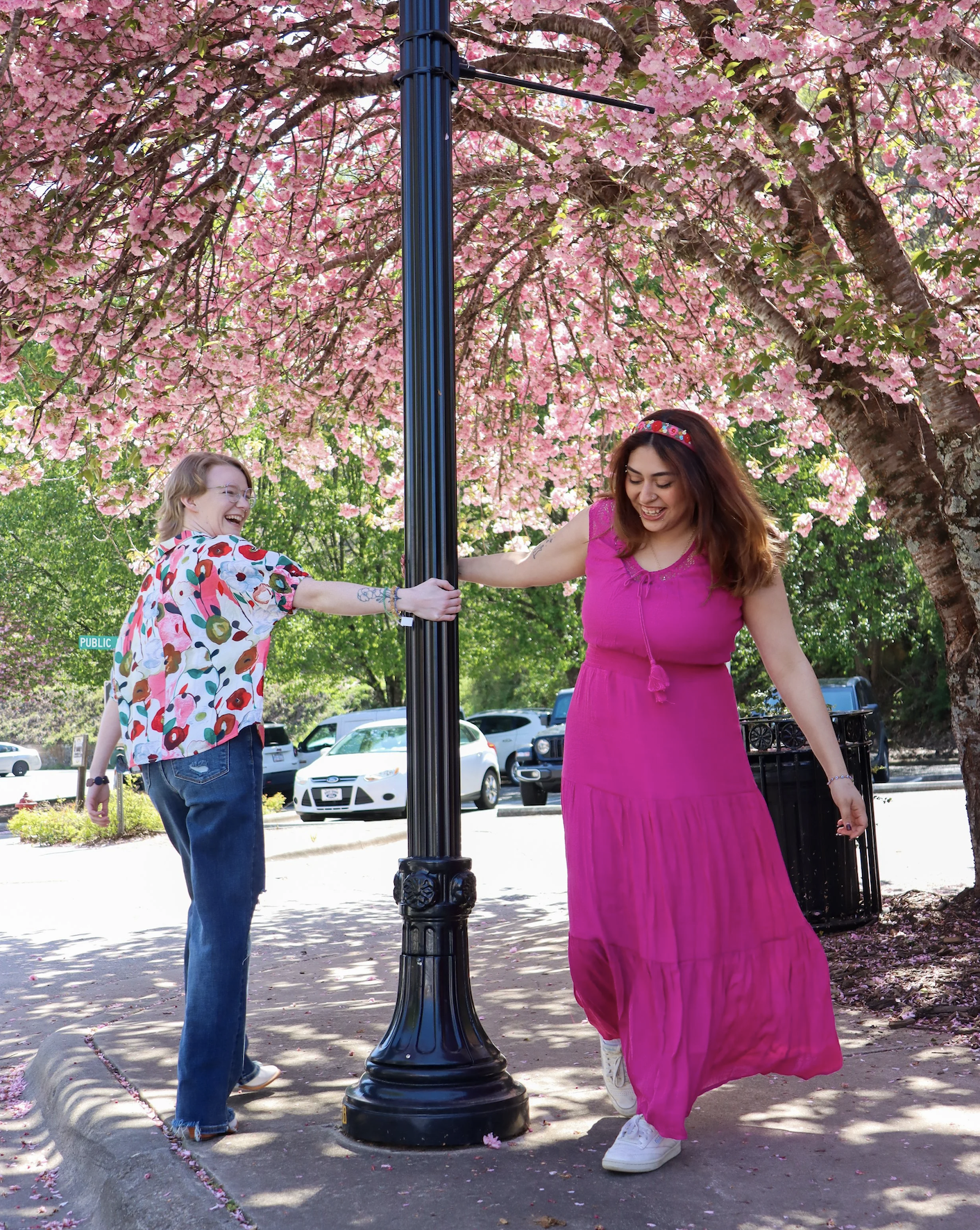 Two women, one in a pink dress and the other in a floral shirt, are playing around a street lamp under a blooming pink cherry blossom tree, smiling and laughing.
