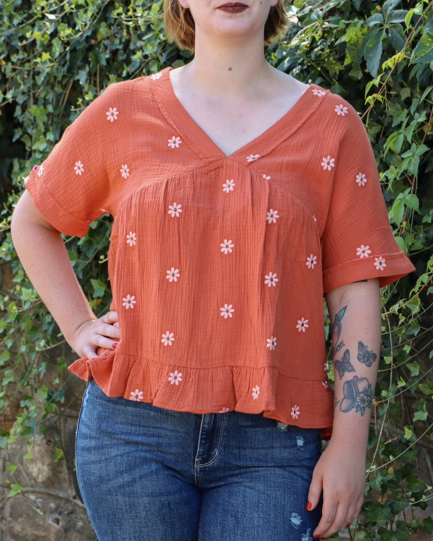 Person wearing an orange top with pink flower patterns, standing outdoors with green foliage in the background.