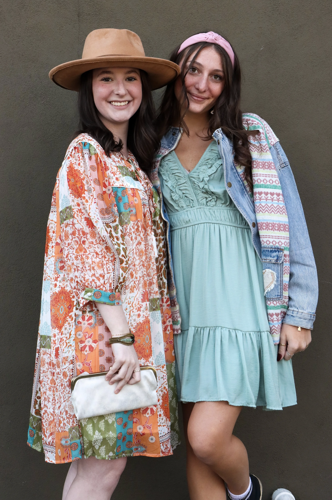 Two young women smiling, standing close against a plain wall. One is wearing a large tan hat and a colorful patterned dress, holding a small clutch. The other is wearing a pink headband, a denim jacket with patterned fabric on the sleeves, and a turq