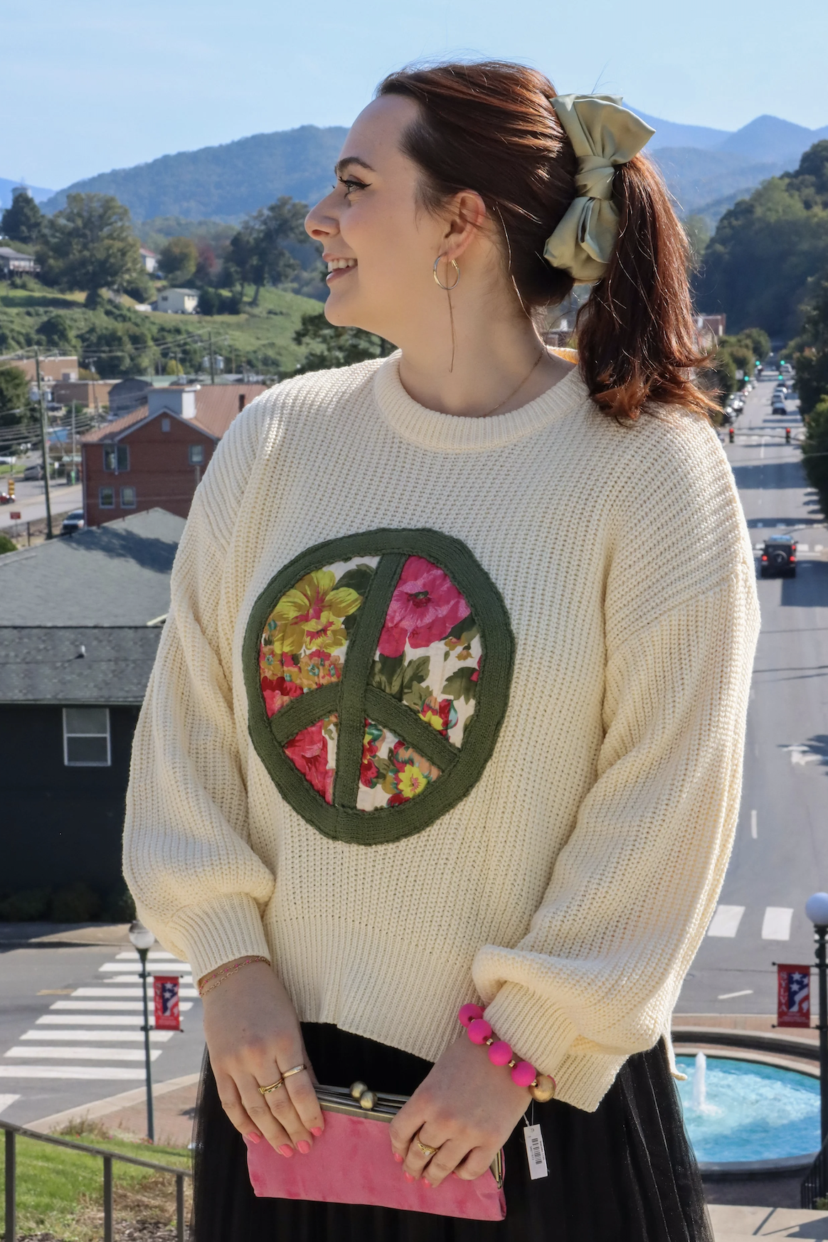 A woman with red hair tied with a beige scrunchie, wearing a cream sweater with a peace symbol filled with floral fabric, smiling and standing outdoors in a town with mountains in the background.