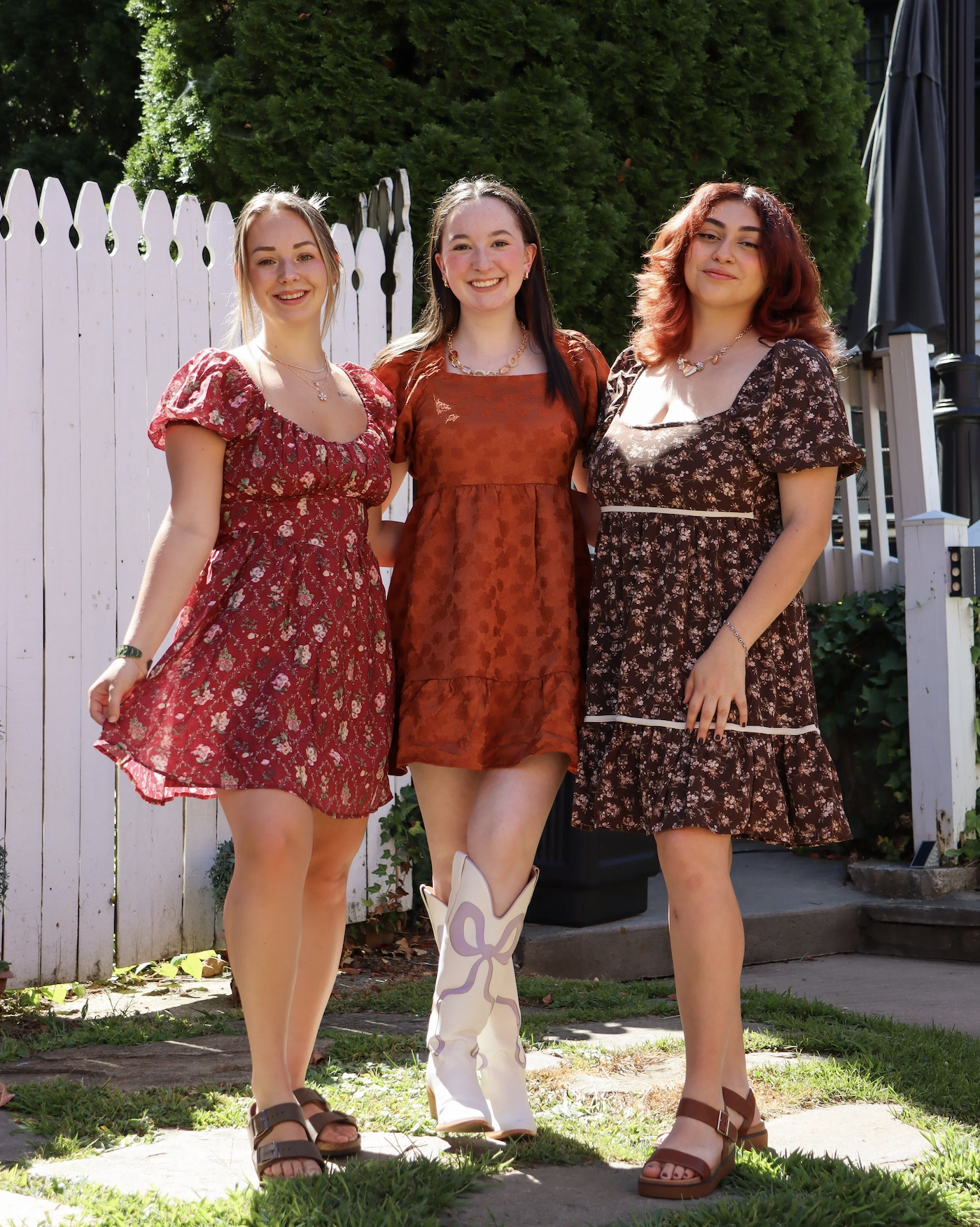 Three young women standing together outdoors near a white picket fence, smiling, in summer dresses.