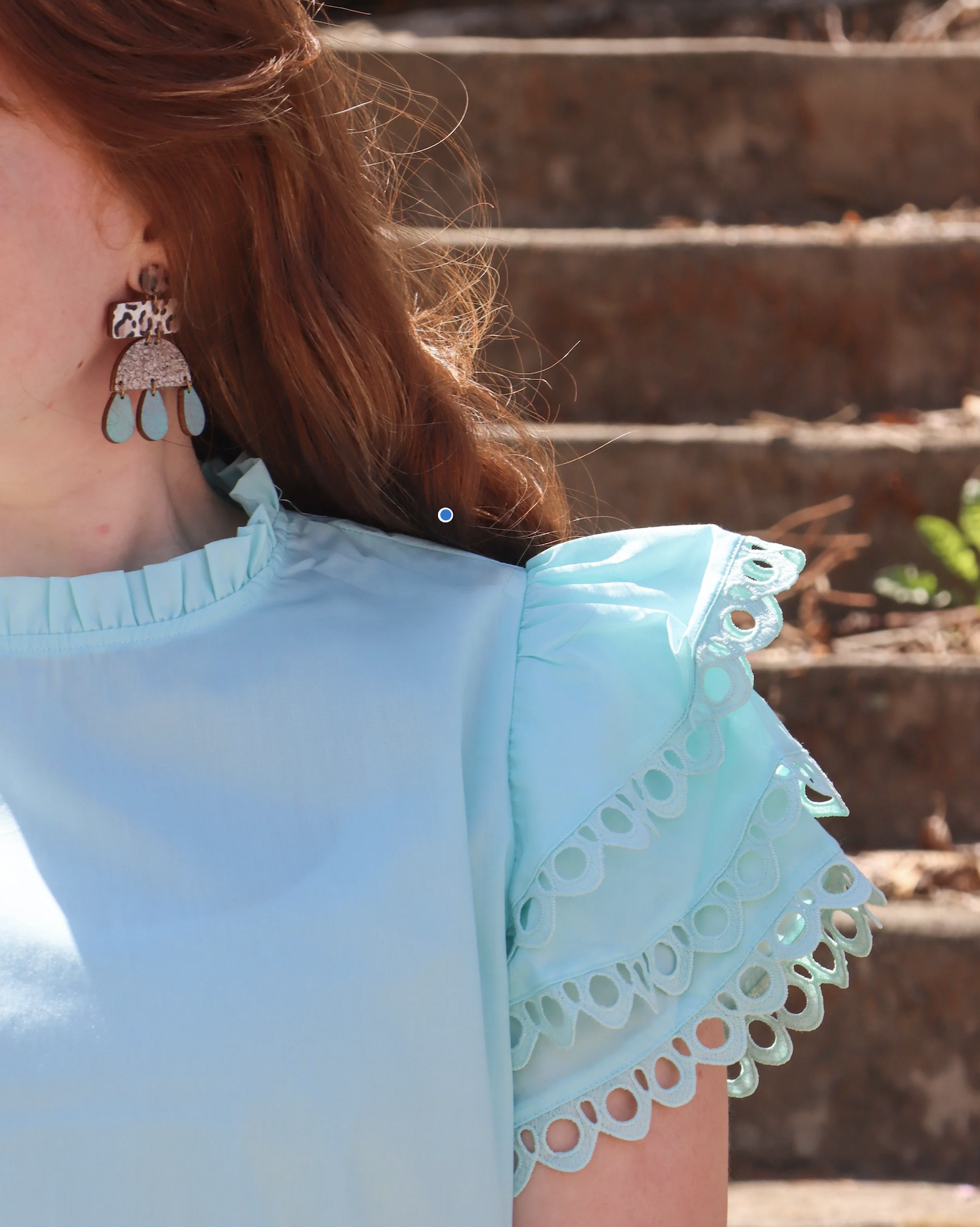 Close-up of a woman wearing turquoise earrings and a light blue top with eyelet lace ruffle sleeves, outdoors near wooden steps.