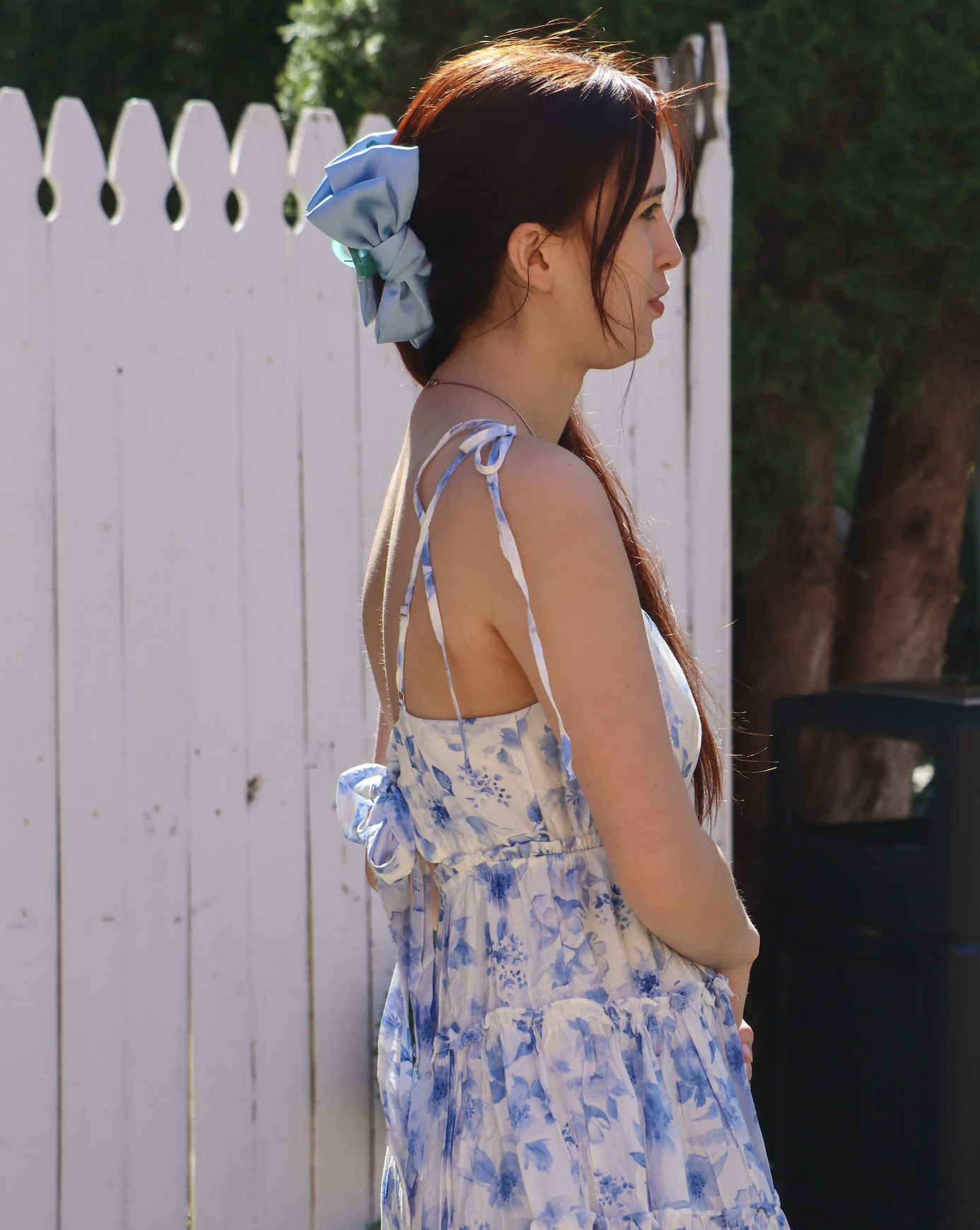 Side profile of a young woman with long reddish-brown hair, wearing a blue and white floral sundress with thin strings on her shoulders, standing outdoors in front of a white picket fence and green trees.