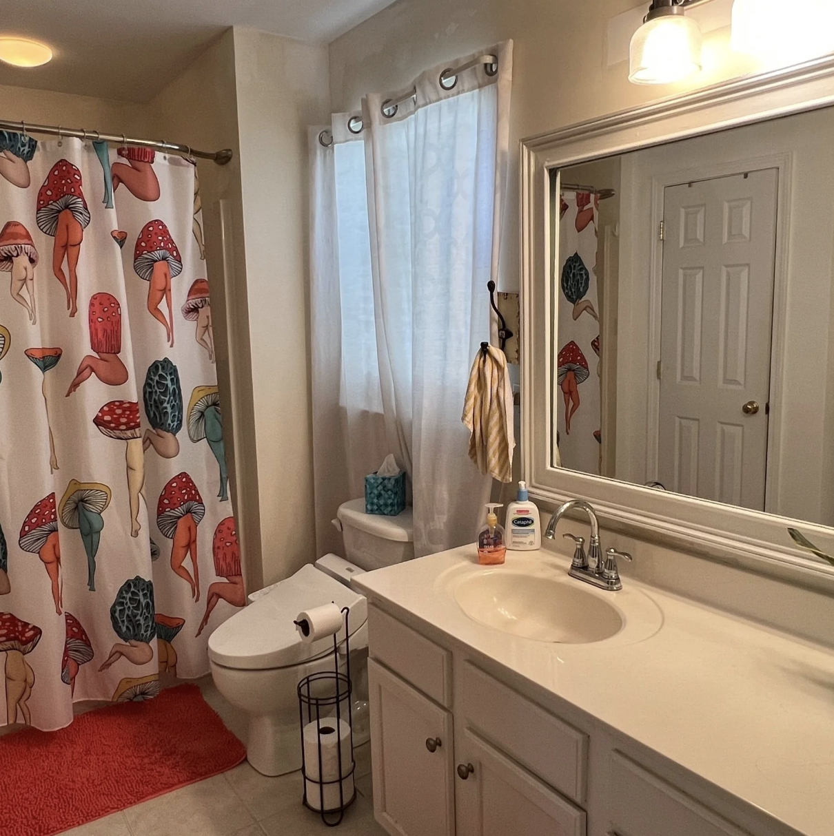 A bathroom with a mushroom-themed shower curtain, a white vanity with a mirror overhead, and a toilet with a tissue holder. There is a window with white curtains and a light fixture above the mirror.