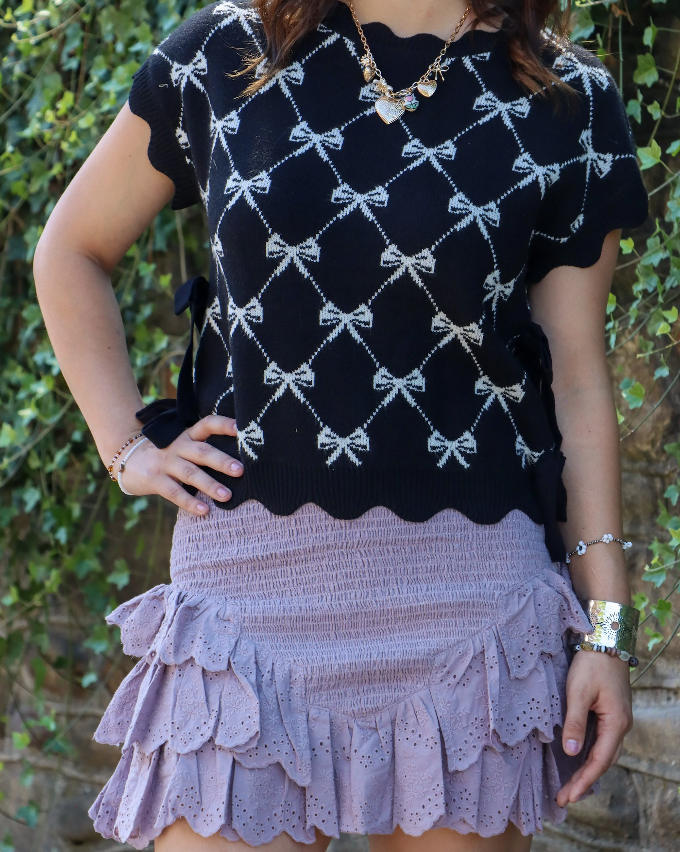 Person wearing a black short-sleeve sweater with a white bow pattern, a pink ruffled skirt with eyelet detailing, and various jewelry, standing outdoors with green foliage in the background.