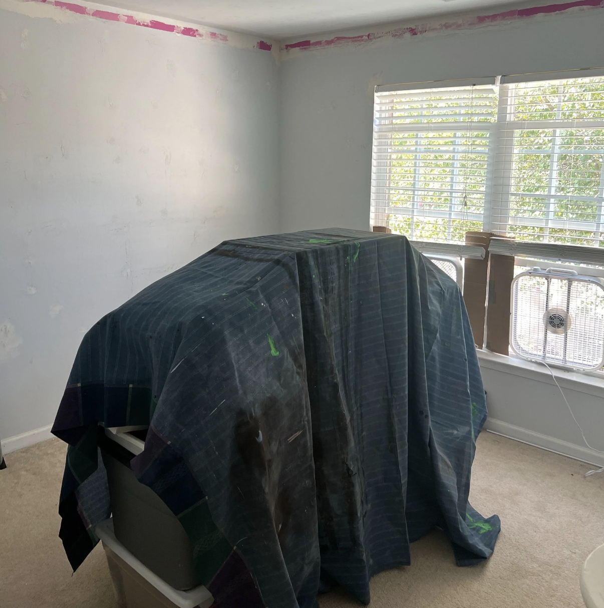 A room undergoing renovation with walls partially painted white, exposed pink paint at the ceiling edges, a window with blinds, and a piece of furniture covered with a dark plaid sheet, with a window fan on the windowsill.