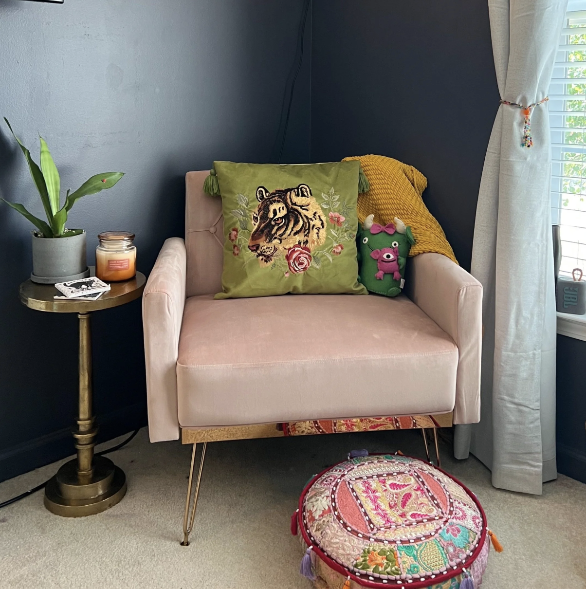 A cozy corner with a pink velvet armchair, decorated with a green throw pillow with a tiger embroidery, a small stuffed pig toy, and a yellow textured blanket. Next to the armchair is a small round brass side table with a potted green plant, a candle