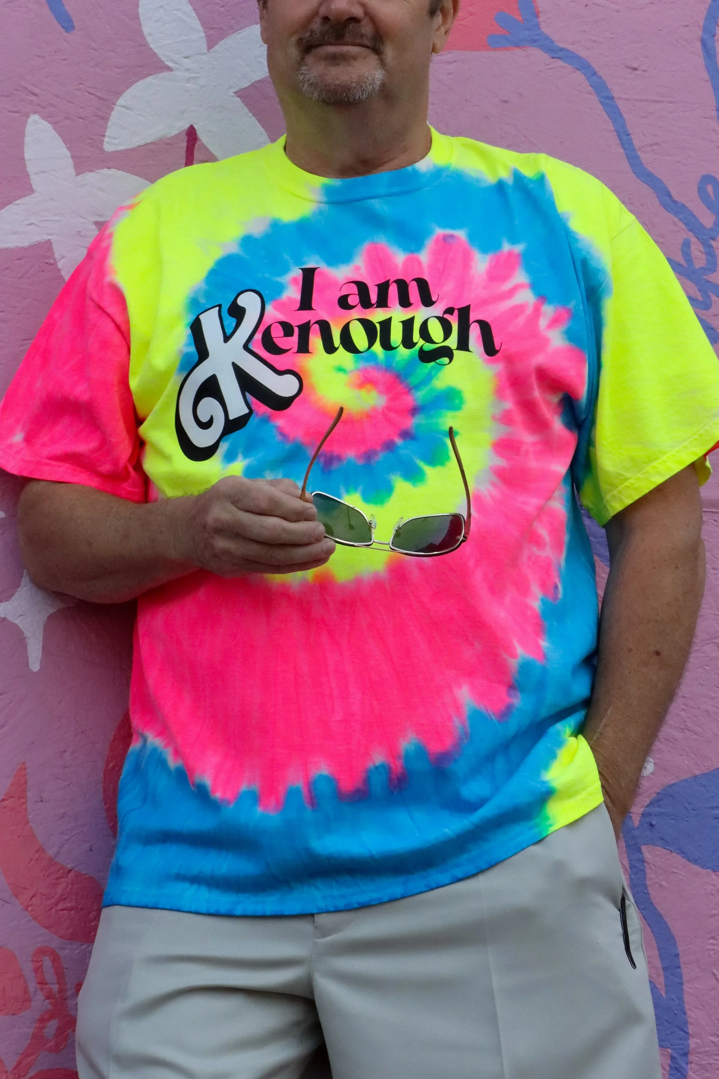 A man wearing a colorful tie-dye shirt with the text "I am X Knough" and holding sunglasses, standing in front of a pink and purple decorative wall.