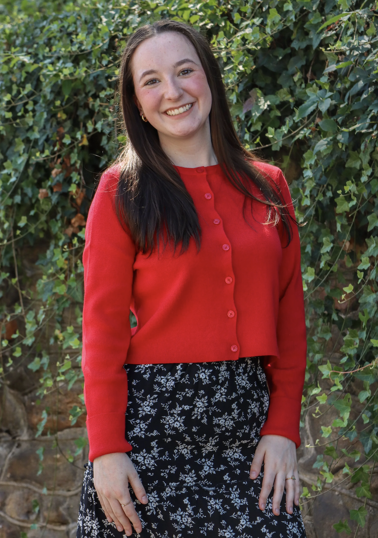 A young woman with long dark hair, smiling, wearing a red cardigan and a black floral skirt, stands outdoors in front of green foliage.