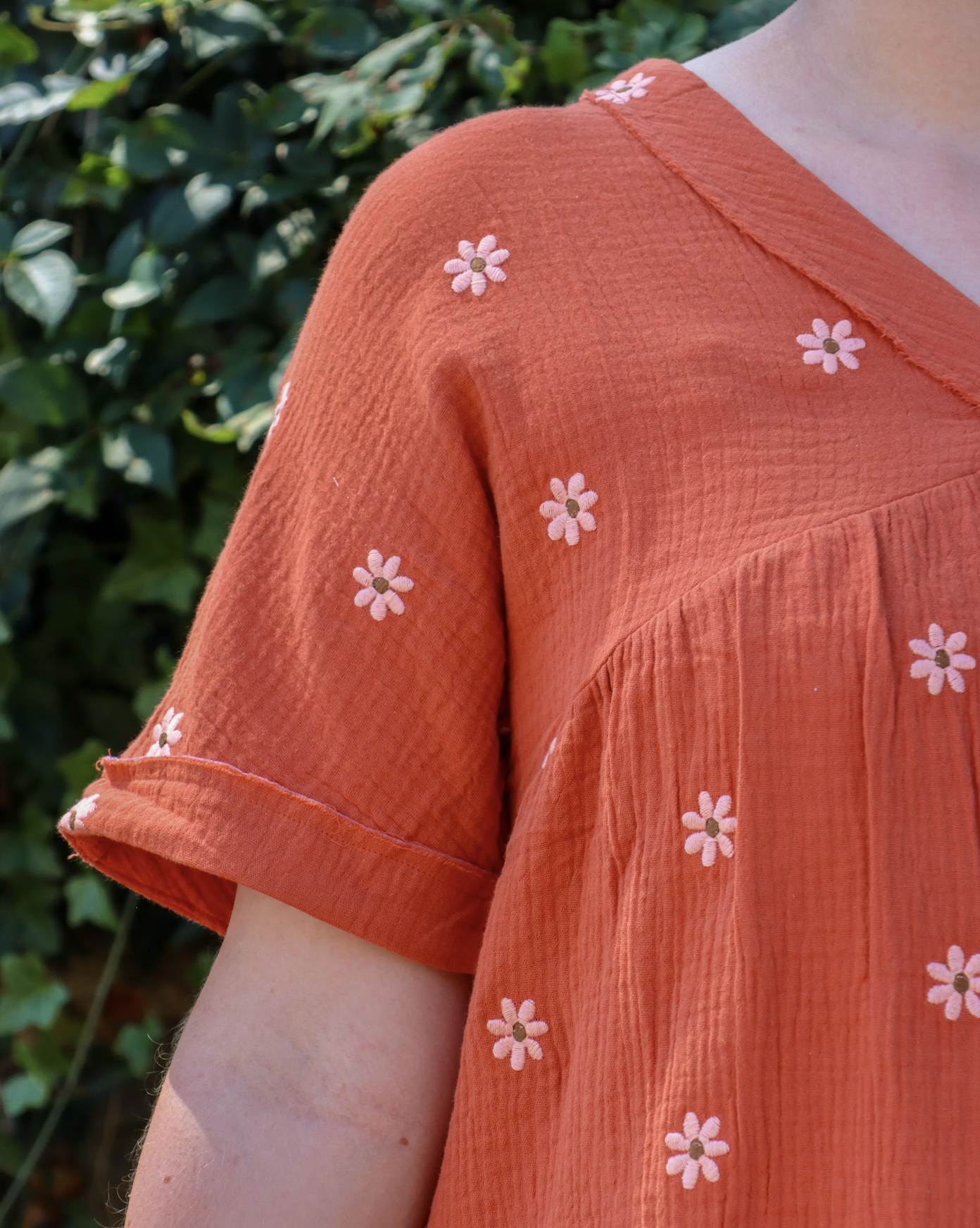 Close-up of a person's shoulder wearing an orange shirt with small embroidered pink and white daisies, standing outdoors with green foliage in the background.