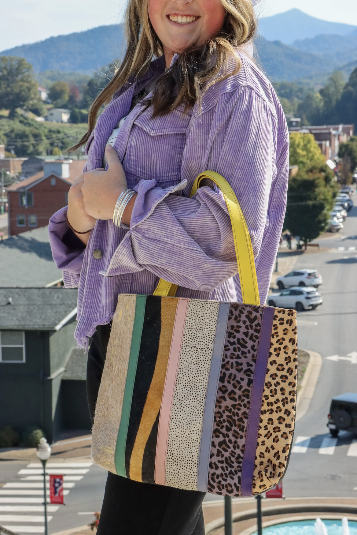 A woman smiling outdoors, wearing a purple striped jacket and holding a colorful, striped tote bag with animal print patterns and a yellow handle.