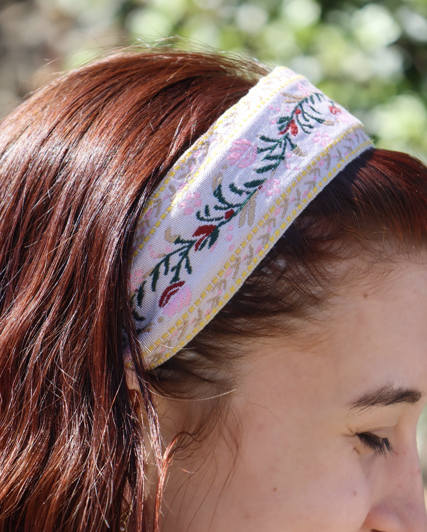 Close-up of a woman with red hair wearing a white headband with colorful floral embroidery, outdoors with blurred greenery in the background.